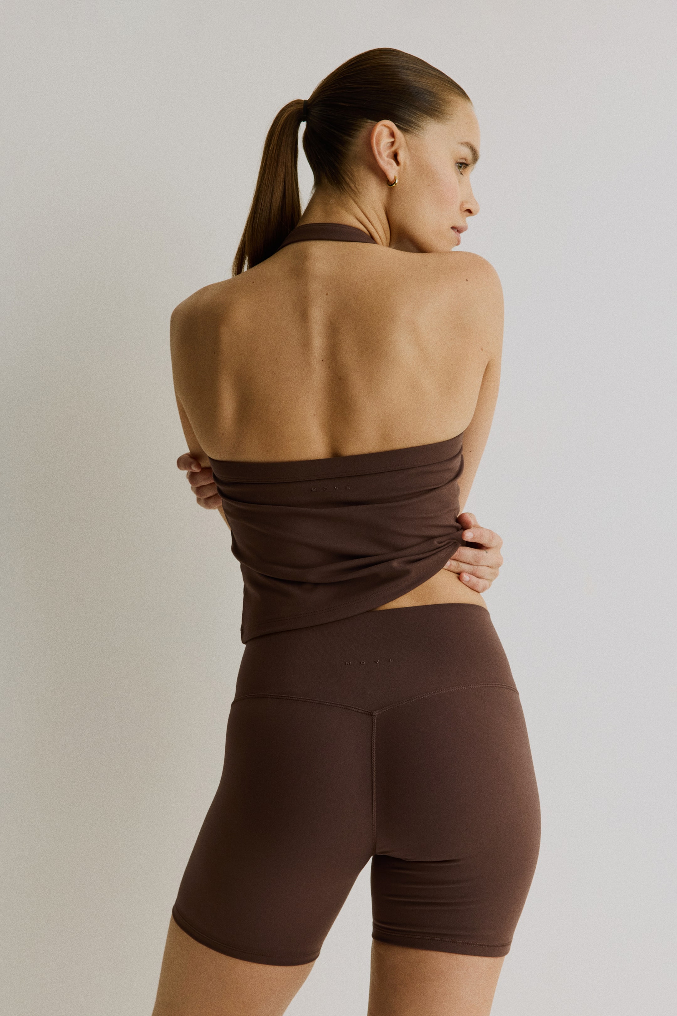 View larger image: The model is wearing a dark brown halterneck sports vest top with SoftMove. The top showcases a visible "MOVE" logo on the back, and the model is holding her lower back with both hands, highlighting its snug fit. She is also wearing matching dark brown sports shorts, which feature the same "MOVE" logo.