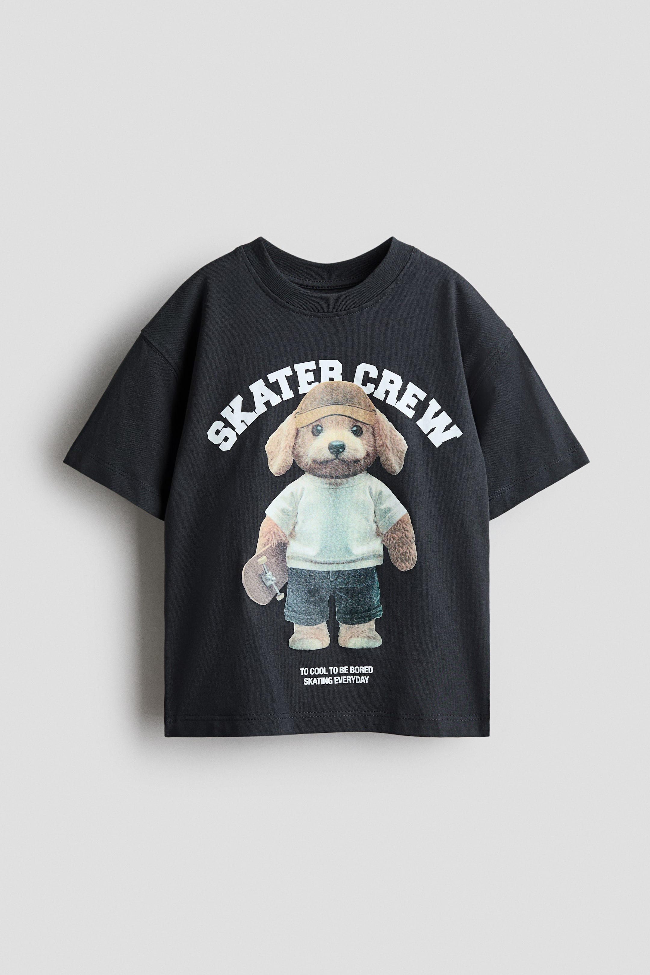 Größeres Bild anzeigen: Ein schwarzes T-Shirt zeigt einen großen Grafikdruck auf der Vorderseite. Dieser Druck zeigt eine Cartoonfigur eines braunen Hundes, der ein helles T-Shirt, blaue Shorts und eine braune Kappe trägt und ein Skateboard hält. Über der Figur steht in weißen Großbuchstaben "SKATER CREW", und darunter in kleinerem Text "TO COOL TO BE BORED SKATING EVERYDAY".