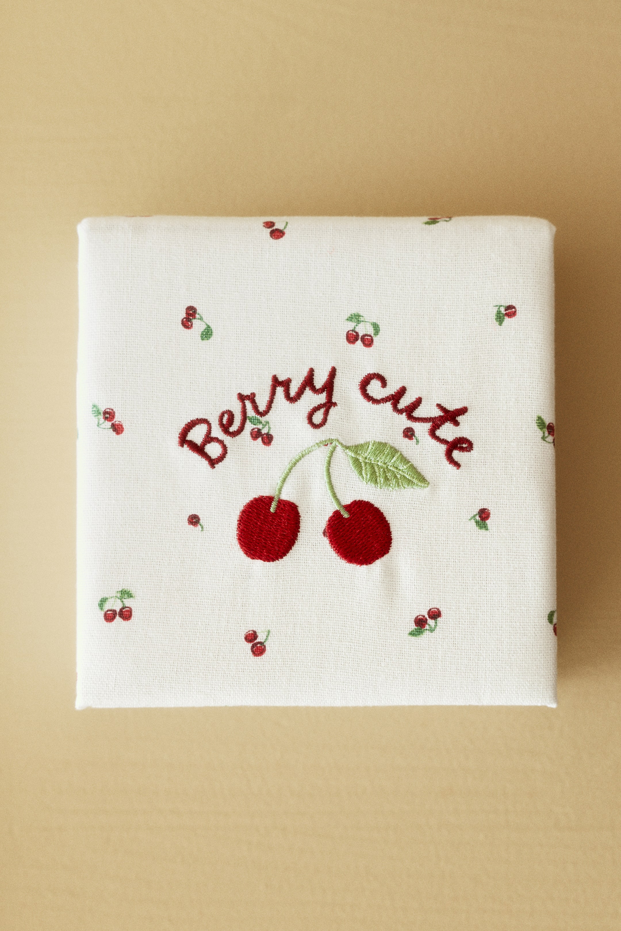 View larger image: A white fabric square is lying flat, showcasing an embroidered design with two large red cherries adorned with green stems and leaves. Above the cherries, the words "Berry cute" are embroidered in a reddish-brown script, while smaller red cherry motifs are scattered across the white fabric.