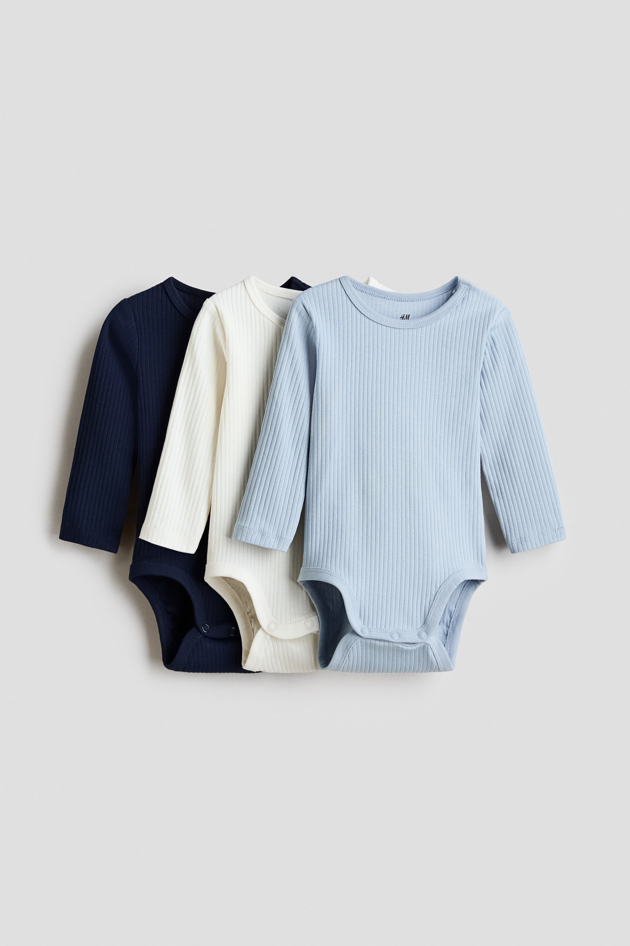 Grotere afbeelding bekijken: Drie babyrompertjes met lange mouwen worden gepresenteerd in lagen, iets naar rechts gekanteld. De rompertjes, in marineblauw, gebroken wit en lichtblauw, hebben een geribbelde breistructuur en drukknoopjes bij het kruis.