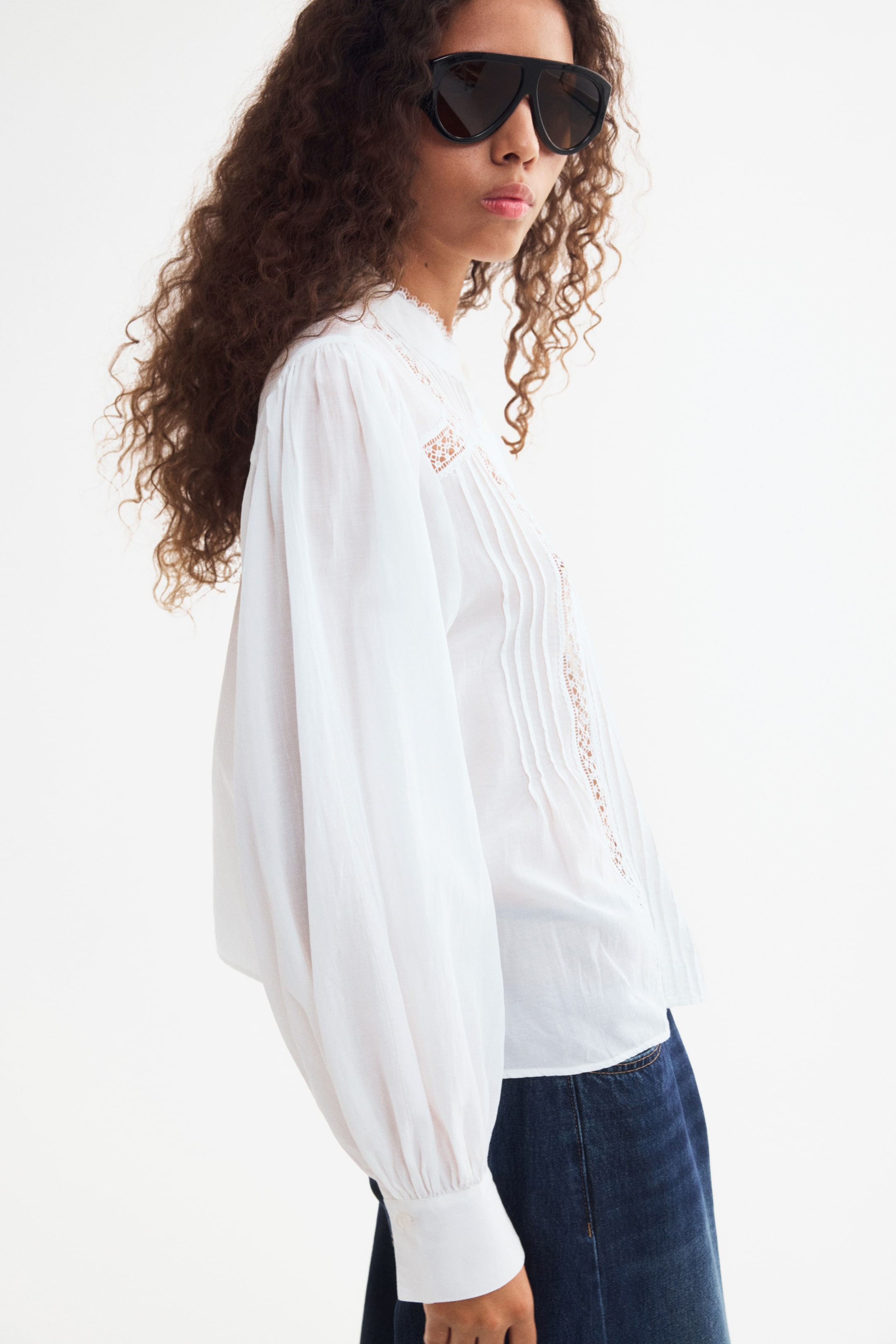 Agrandir l'image: Le mannequin porte une blouse blanche à la coupe ample, associée à un jean bleu foncé. La blouse présente un col officier bordé de dentelle, des plis verticaux décoratifs agrémentés d’empiècements en dentelle sur le devant, ainsi que de longues manches volumineuses avec des poignets boutonnés froncés.