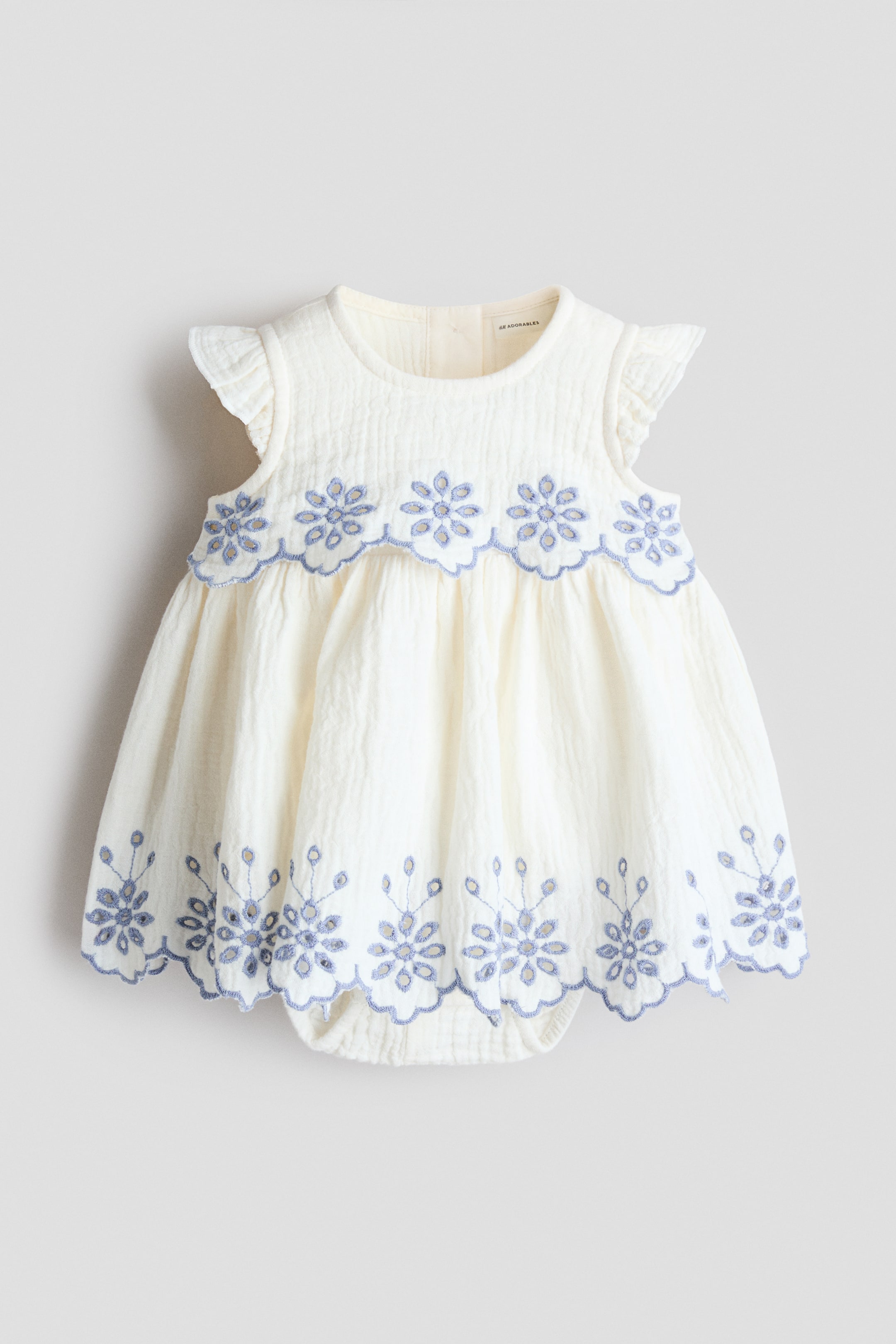 Voir une image plus grande: Une robe crème pour nourrisson présente un tissu texturé et froissé avec une broderie florale bleue, vue de face. Elle est conçue avec un col rond, des manches à volants et une jupe froncée, et affiche une bande brodée festonnée sous le col et une autre près de l'ourlet. La robe intègre également un bas de style barboteuse avec des ouvertures de jambes élastiquées.