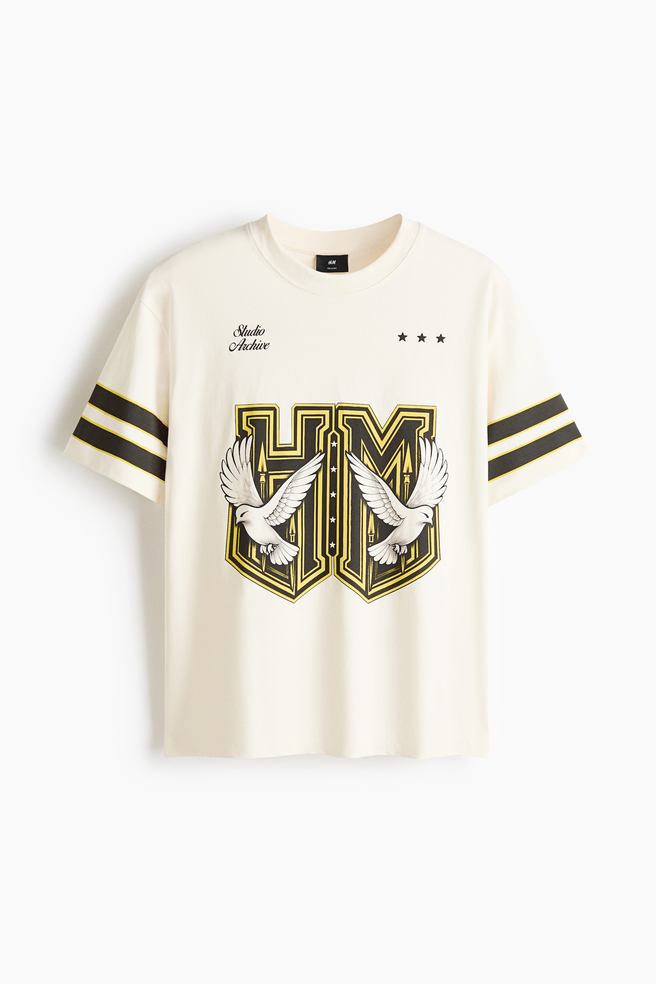 View larger image: A cream t-shirt is displayed flat, facing forward. It features a round neck and short sleeves adorned with two black and one yellow stripe on each. A large central graphic showcases yellow and black 'HM' lettering flanked by two white doves. The chest area also displays 'Studio Archive' text and three black stars.