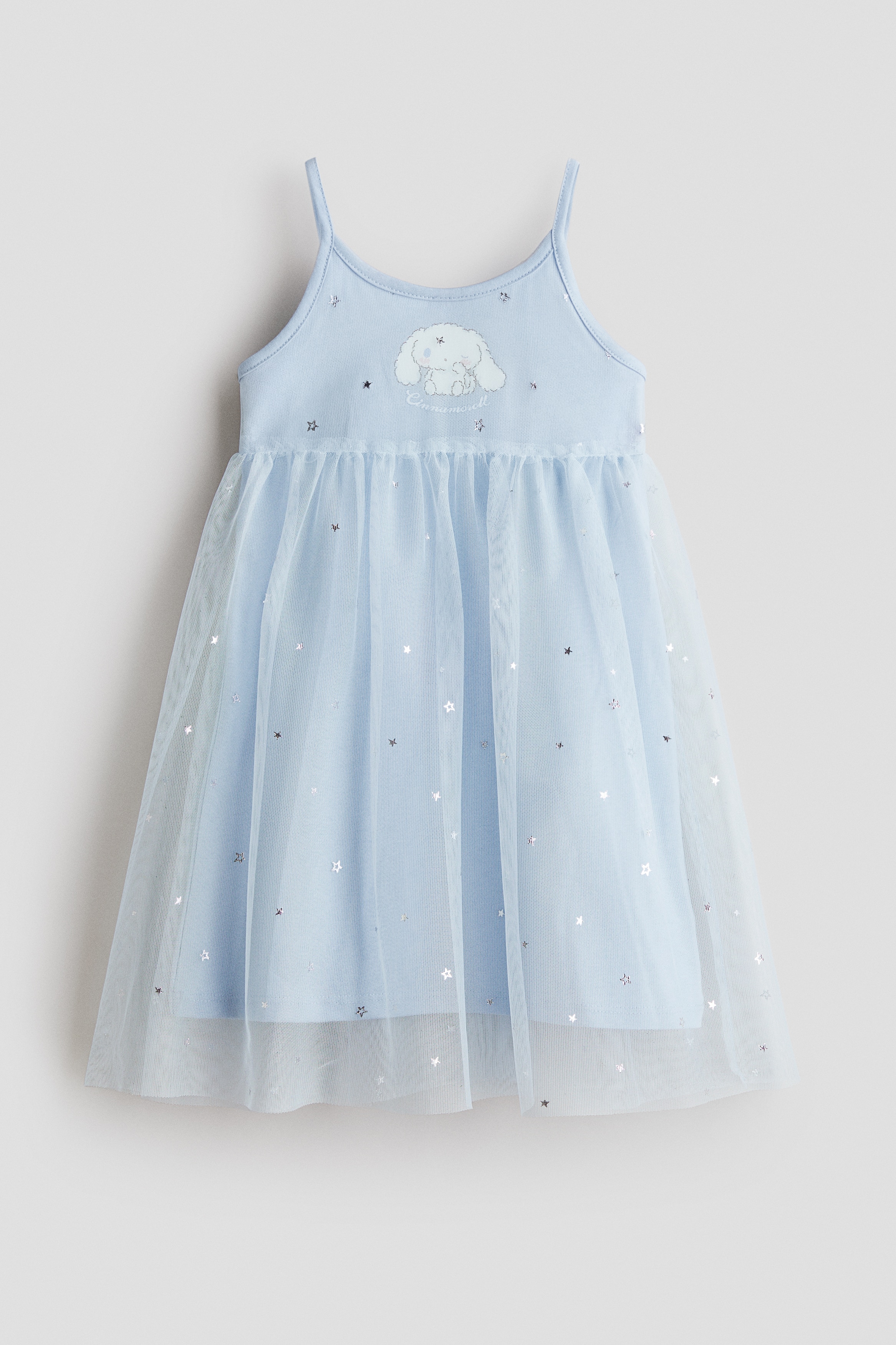 A light blue jersey dress with narrow shoulder straps and a printed character on the chest is worn by the model. The dress features a flared skirt overlaid with a sheer, star-patterned tulle layer and is paired with silver metallic flat shoes.
