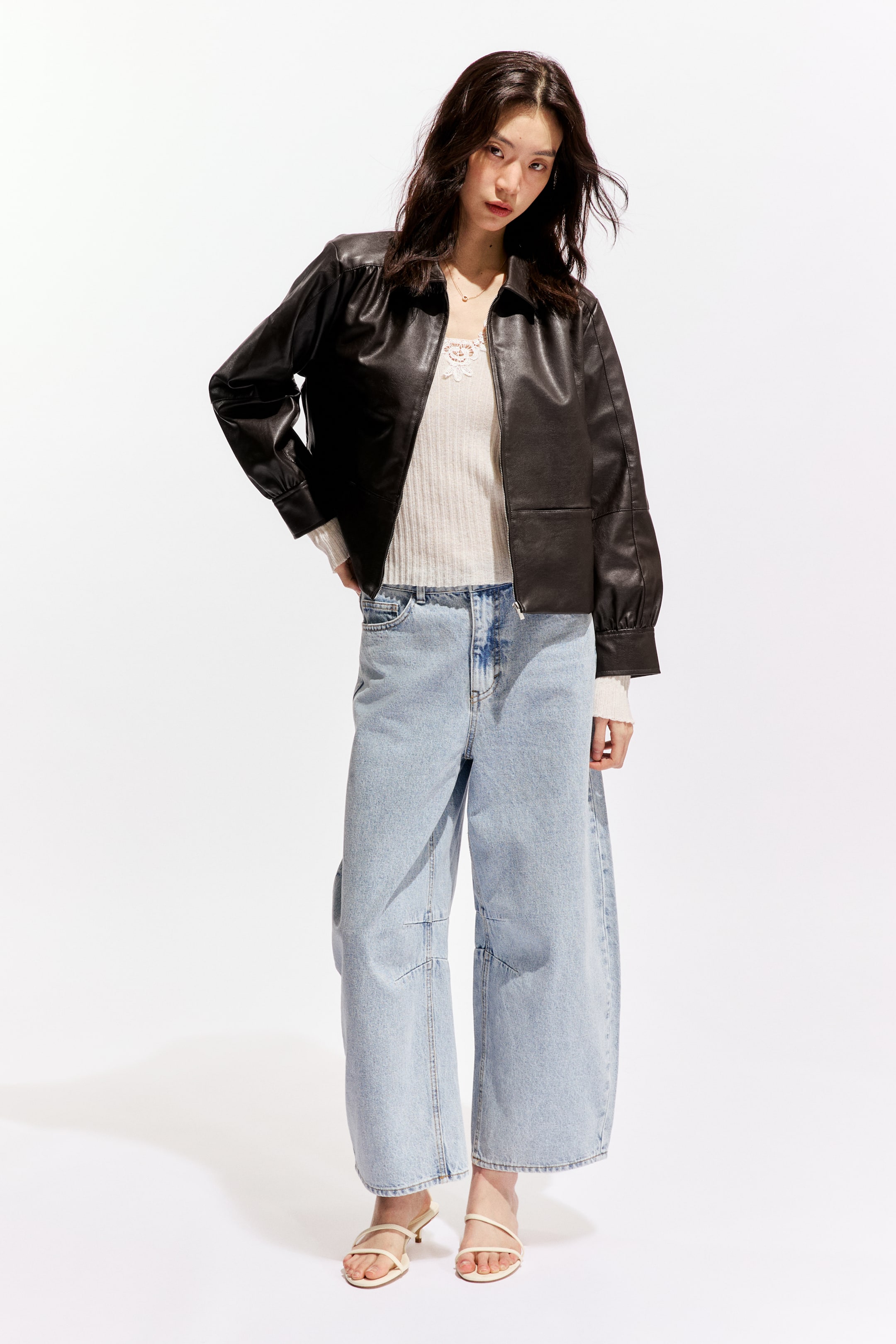 拡大画像を表示: 黒のフェイクレザージャケットが紹介されています。尖った襟、肩にわずかなボリュームのある長袖、ギャザー入りの袖口、下前部に長方形の縫い目のディテールがあり、白いリブトップの上にオープンな状態で着用されています。