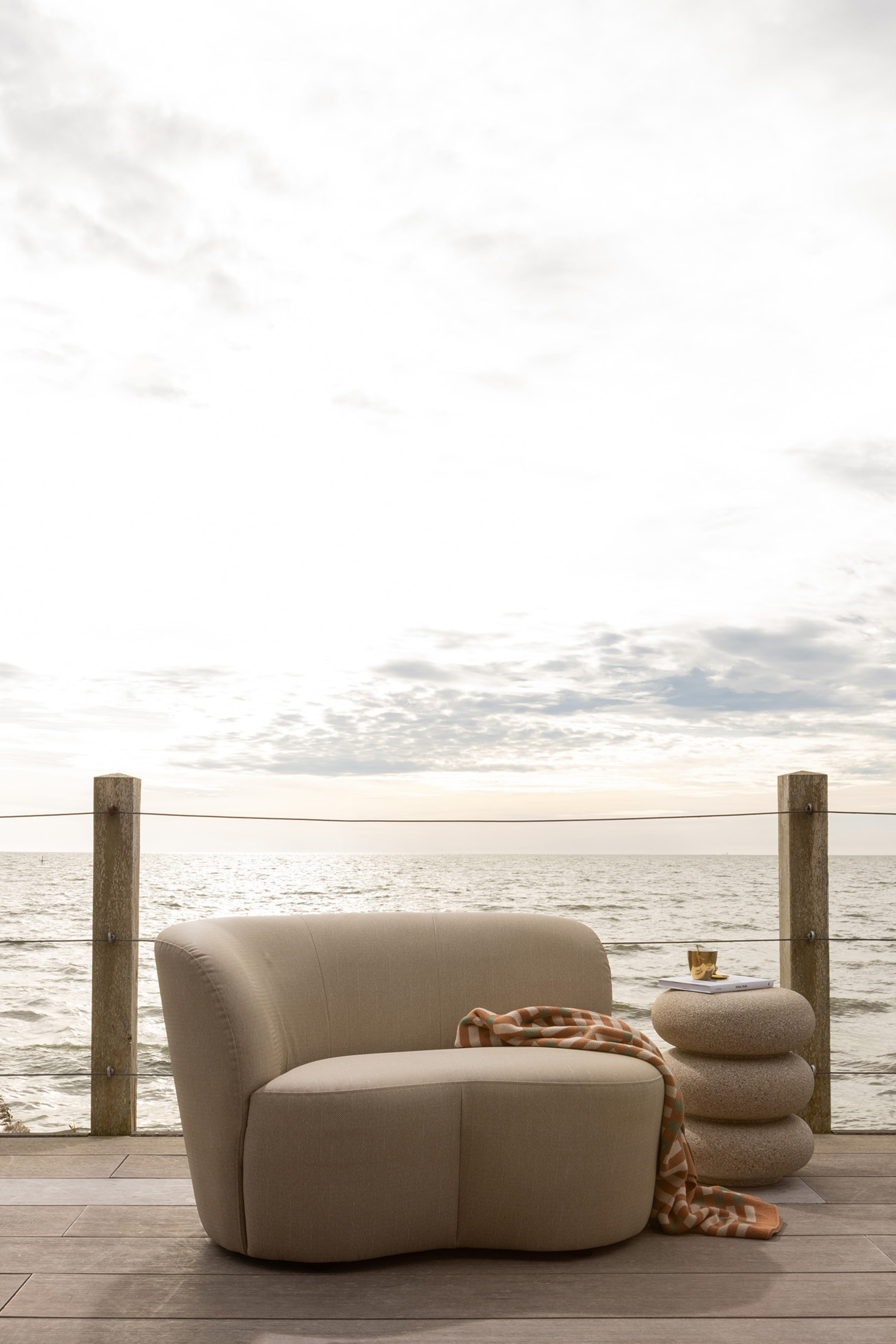 Agrandir l'image: Un fauteuil de salon beige clair présente sa silhouette arrondie avec un dossier et des accoudoirs intégrés, tandis qu'une couverture rayée est drapée sur son siège et son côté.