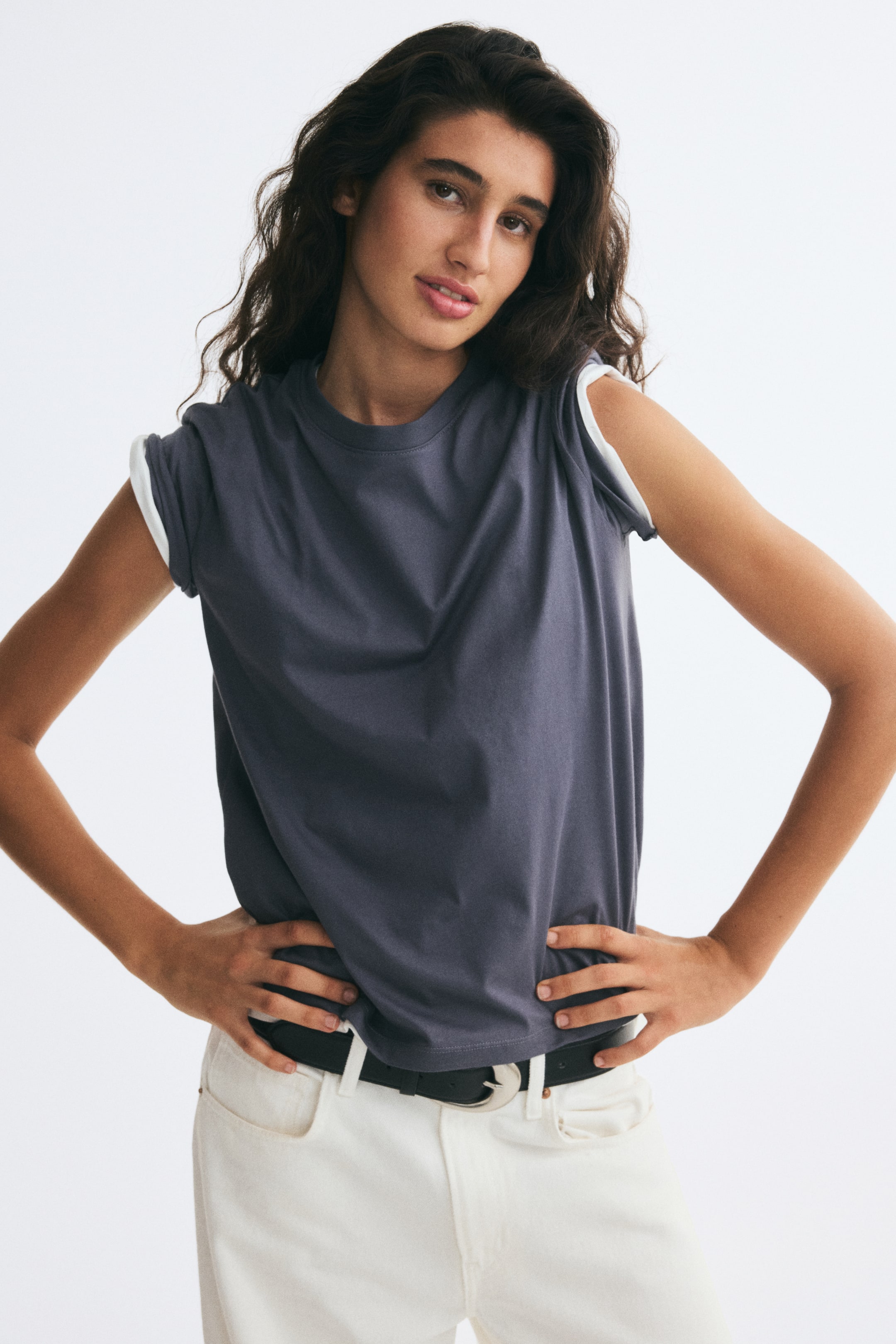 Ampliar la imagen: La modelo lleva una playera de corte recto en gris oscuro, confeccionada en suave jersey de algodón, con escote redondo ribeteado y hombros ligeramente caídos con mangas remangadas que dejan ver una prenda blanca en capas debajo. La playera se combina con jeans blancos y un cinturón negro.