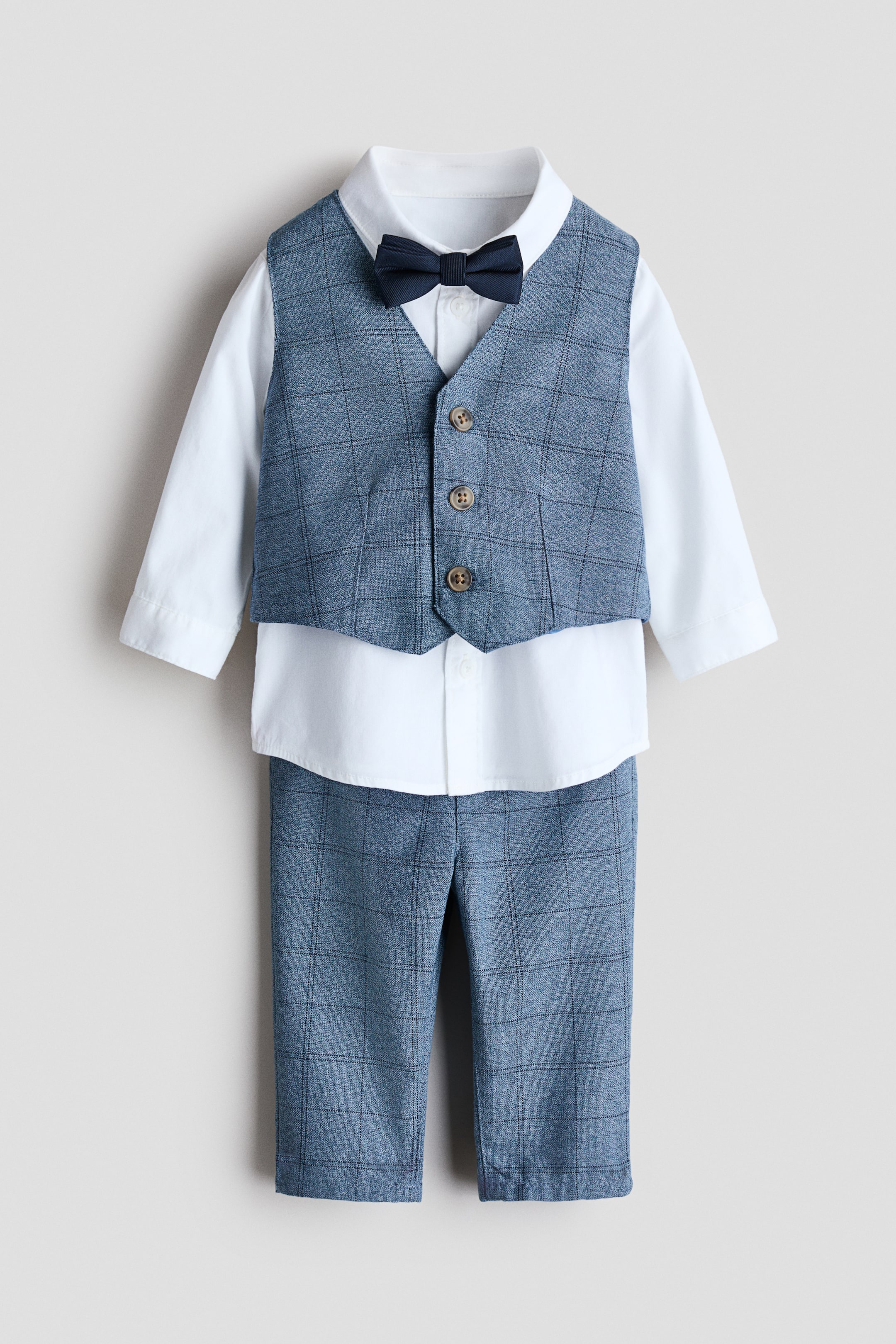 Visualizza l'immagine ingrandita: Un set ben vestito presenta un gilet e pantaloni con motivo blu-grigio, una camicia bianca a maniche lunghe e un papillon blu navy, tutti rivolti in avanti. Il gilet presenta tre bottoni marroni sul davanti ed è sovrapposto alla camicia bianca, che mostra un colletto con bottoni e polsini abbottonati. I pantaloni presentano una finta patta con bottone, abbinata al motivo del gilet, e il papillon blu navy è attaccato al colletto della camicia.