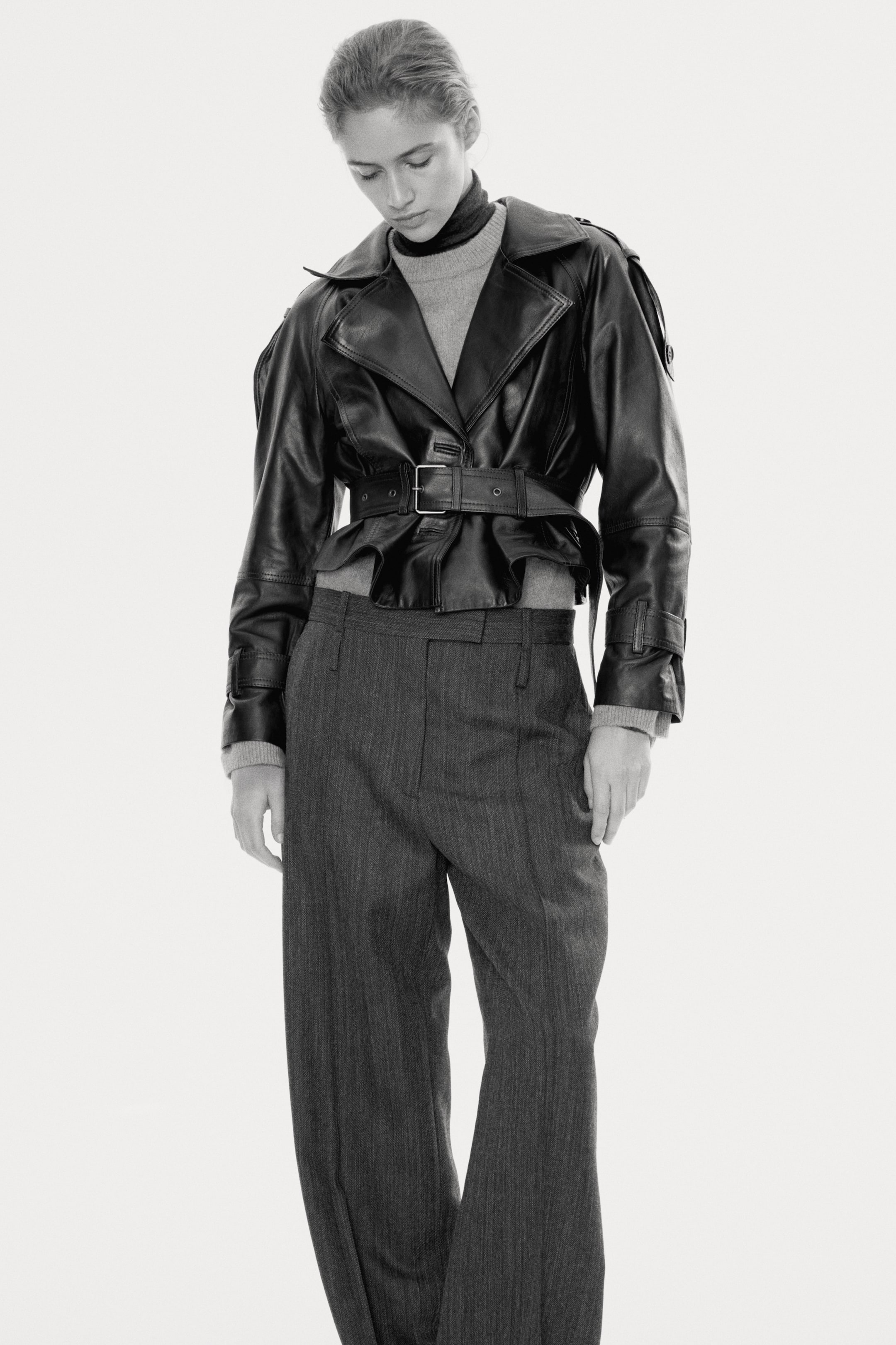 View larger image: A short, double-breasted biker jacket in soft leather is being worn by the model, cinched at the waist with an adjustable belt featuring a metal buckle. The jacket, which features wide notch lapels and buttoned shoulder tabs, is styled over a high-neck knit sweater and wide-leg trousers.