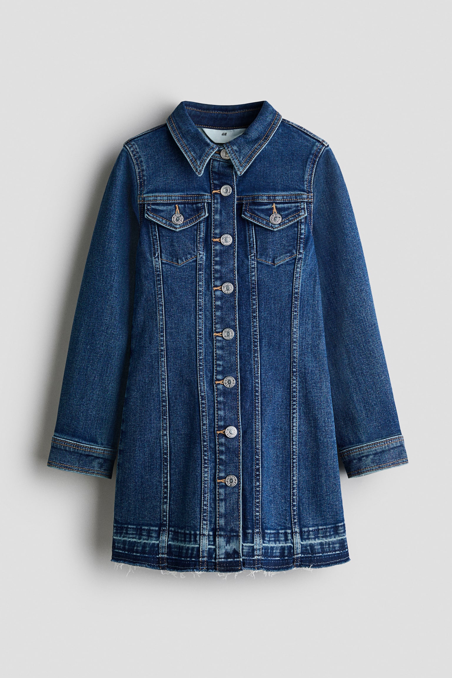 Une robe en denim Ã  manches longues dans un lavage bleu foncÃ© est prÃ©sentÃ©e de face, avec un col pointu, deux poches poitrine Ã  rabat boutonnÃ©es et une fermeture boutonnÃ©e sur toute la longueur avec des boutons mÃ©talliques argentÃ©s. La robe prÃ©sente des dÃ©tails de coutures verticales sur le devant et un ourlet brut lÃ©gÃ¨rement effilochÃ©.