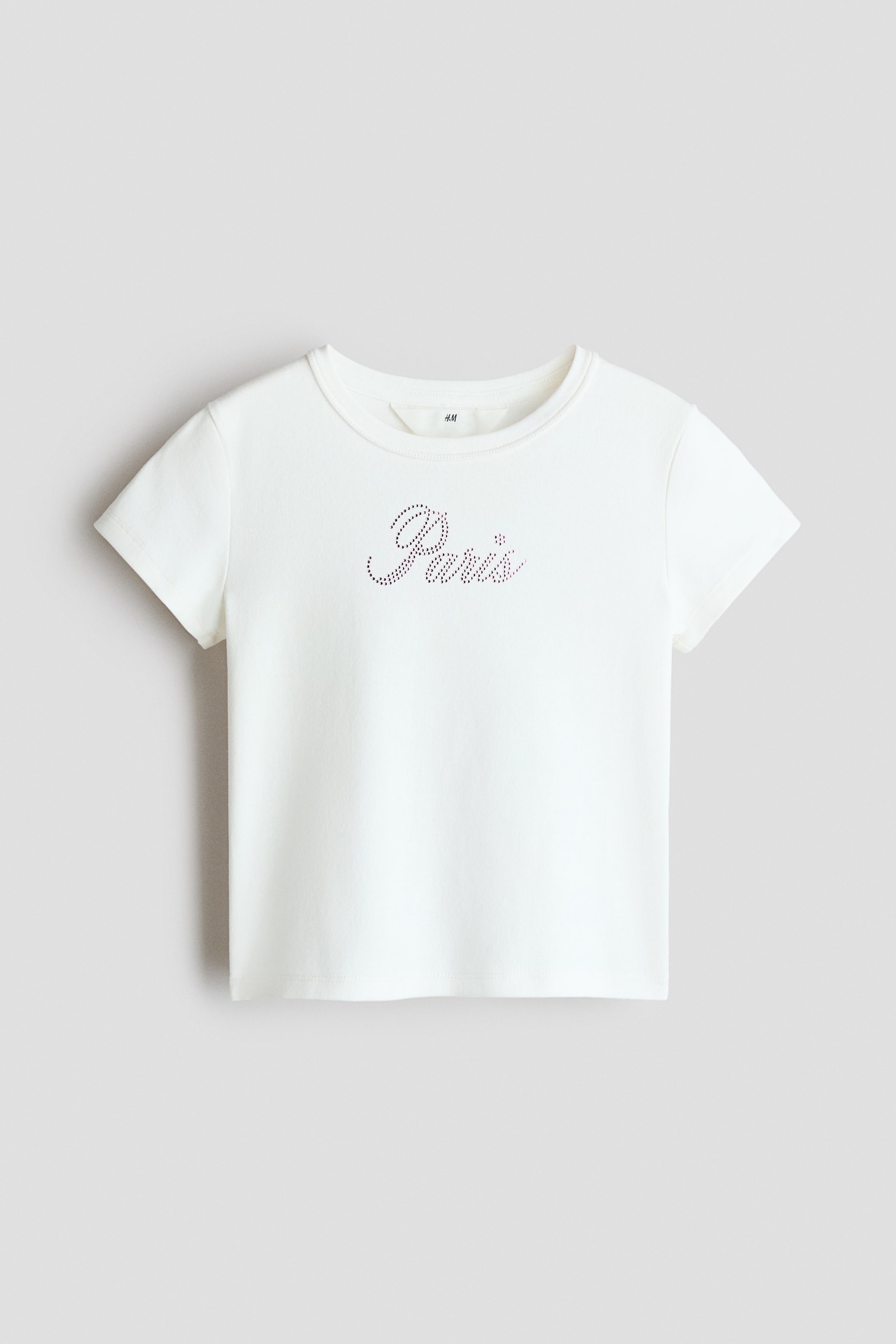 View larger image: A cream-colored, soft cotton jersey tee is facing FRONT, featuring short sleeves and a ribbed trim around the neckline. An embellished "Paris" text motif is visible on the front.