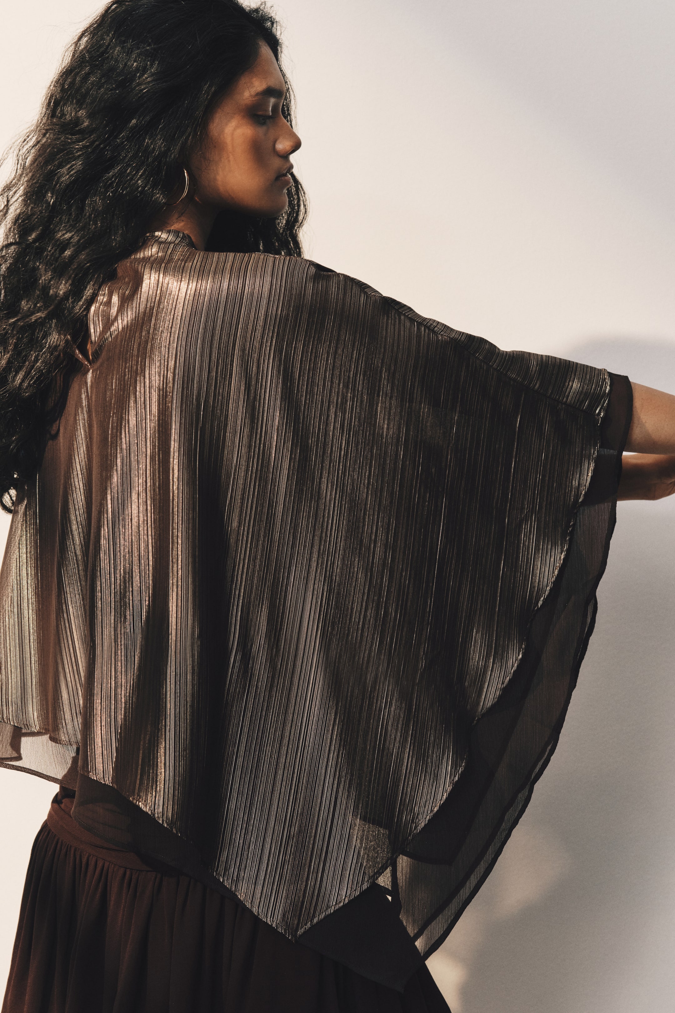 View larger image: A cropped, loose-fit cape in metallic, textured bronze fabric is being worn by the model, who is standing in profile with her arms slightly raised. The cape features a draped silhouette with an asymmetrical handkerchief hem and a sheer lining visible at the edges.