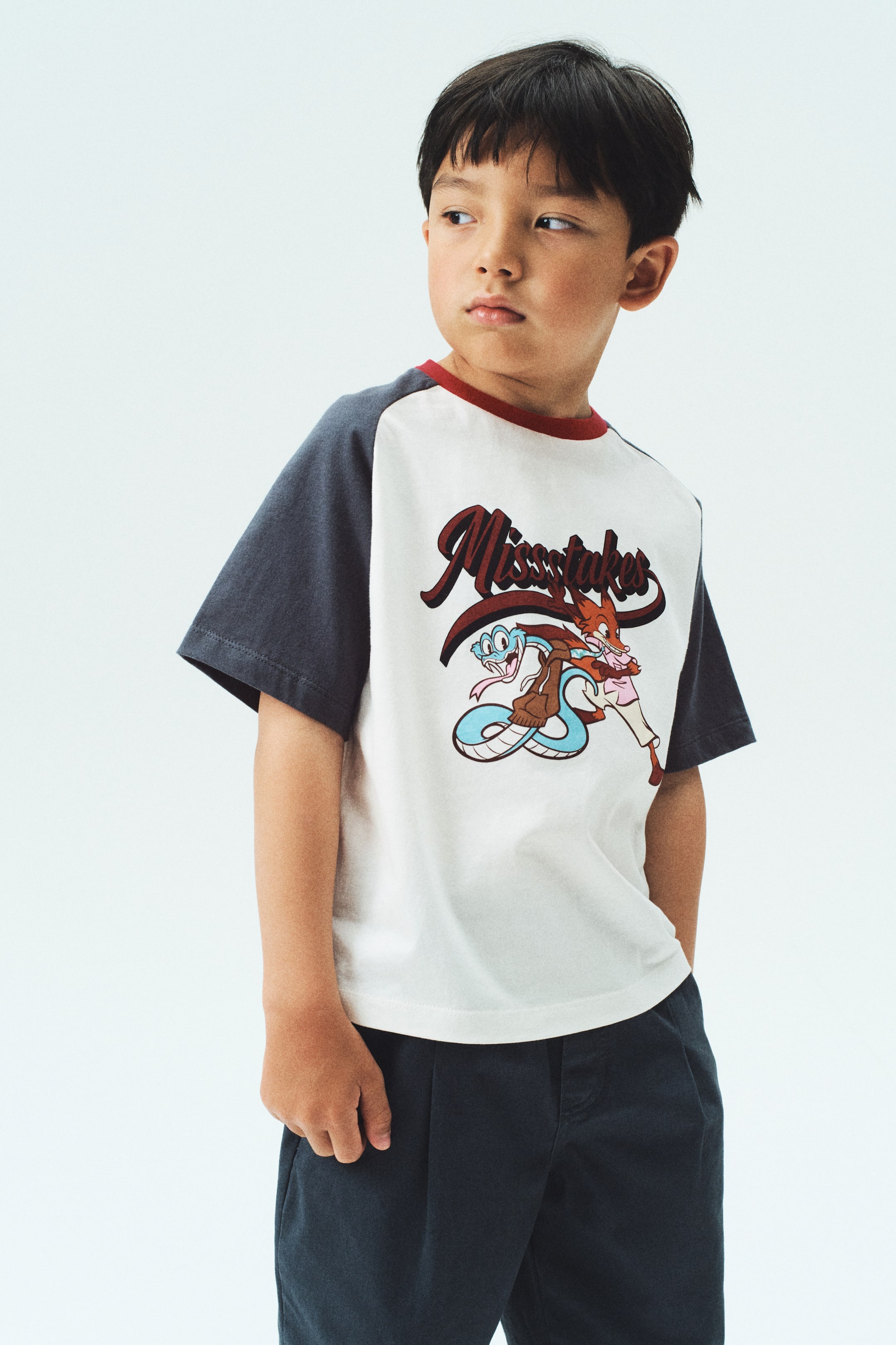 拡大画像を表示: Tシャツは、オフホワイトのボディにダークグレーのラグランスリーブと赤いリブ付きクルーネックを特徴としており、わずかに右前方からの角度で提示されています。フロントには、ダークレッドで「Missstakes」というテキストと、青いヘビと茶色のキツネの漫画キャラクターが描かれています。Tシャツはダークパンツの上に着用されており、そのカジュアルなフィット感と長さを示しています。