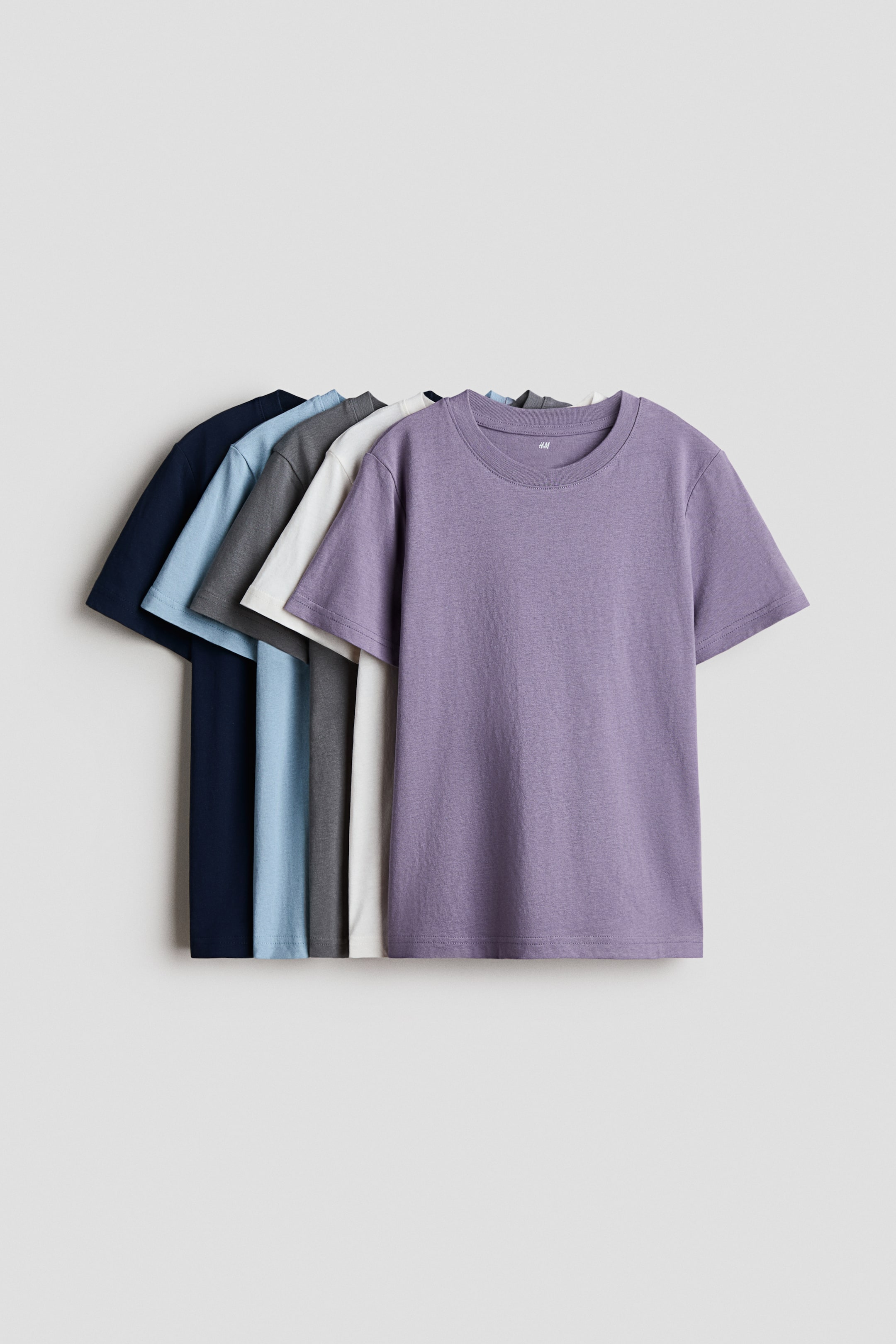 Agrandir l'image: Un ensemble de t-shirts en jersey de coton doux présente des cols ronds et des ourlets droits. Le t-shirt violet à l'avant est exposé en vue complète de face, tandis que les autres t-shirts en bleu marine, bleu clair, gris foncé et blanc cassé se superposent partiellement derrière lui.