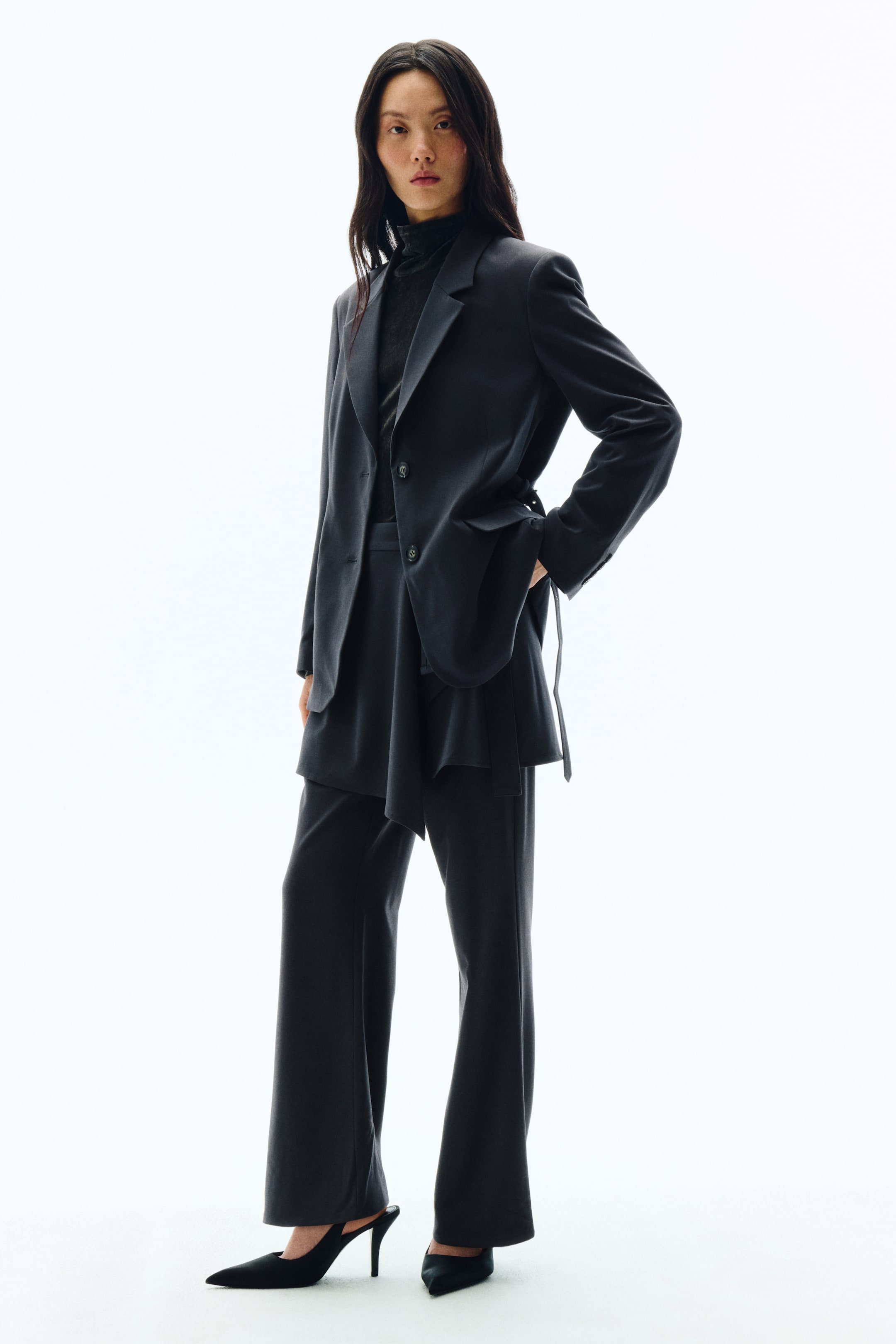 拡大画像を表示: ダークチャコールのスーツが、やや右斜め前からのアングルで撮影されています。オーバーサイズシルエットのシングルブレストブレザーは、ノッチドラペルを備え、サイドタイによってアシンメトリーなドレープのオーバーレイが生まれ、同素材のワイドレッグトラウザーとセットアップでスタイリングされています。インナーにはダークカラーのタートルネックをレイヤードし、足元はブラックのポインテッドトゥ・スリングバックヒールでコーディネートを完成させています。