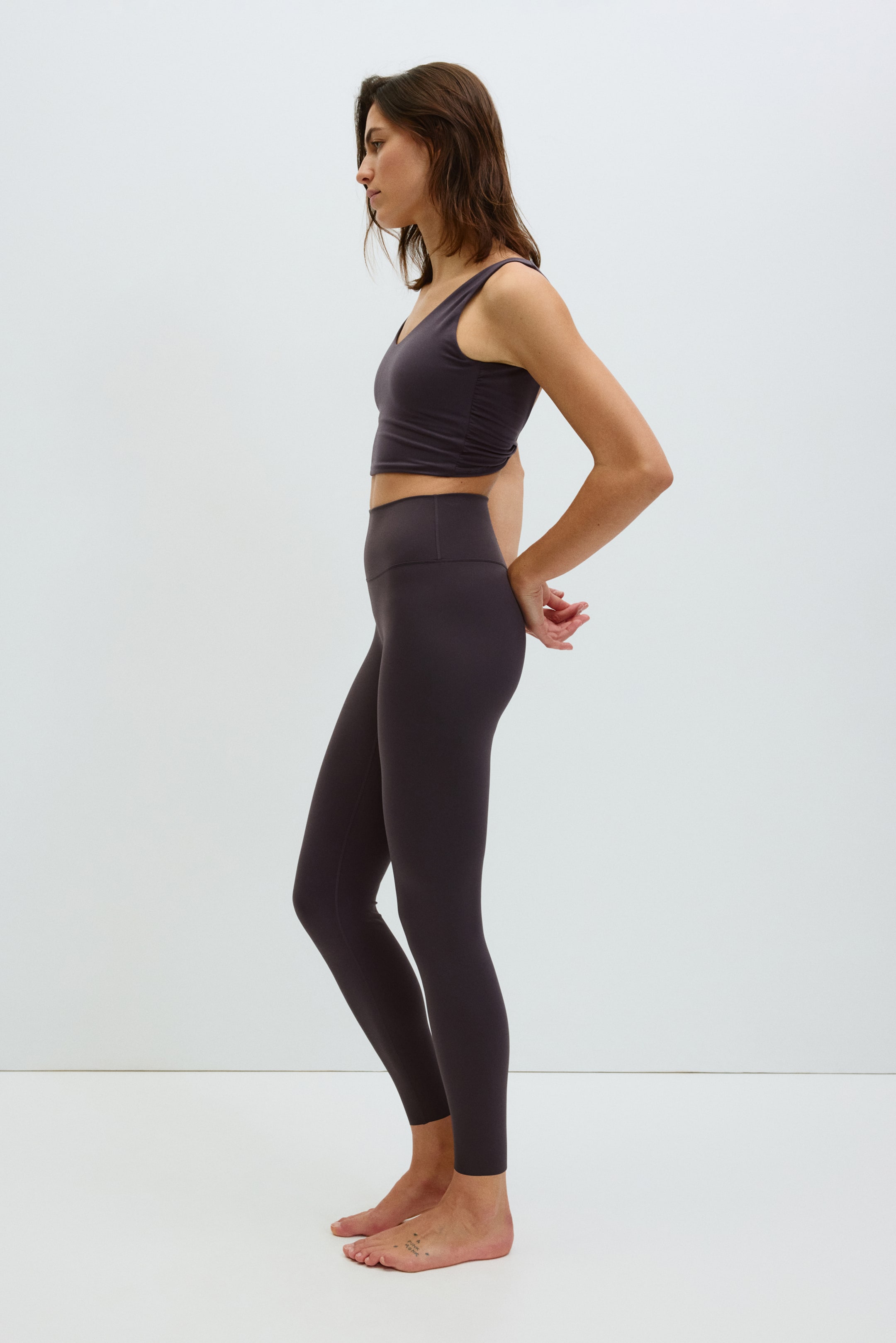 View larger image: A dark grey athletic crop top and matching high-waisted leggings are being shown from a side angle. The crop top features a V-neckline and side ruching, while the leggings present a smooth, form-fitting design down to the ankles.