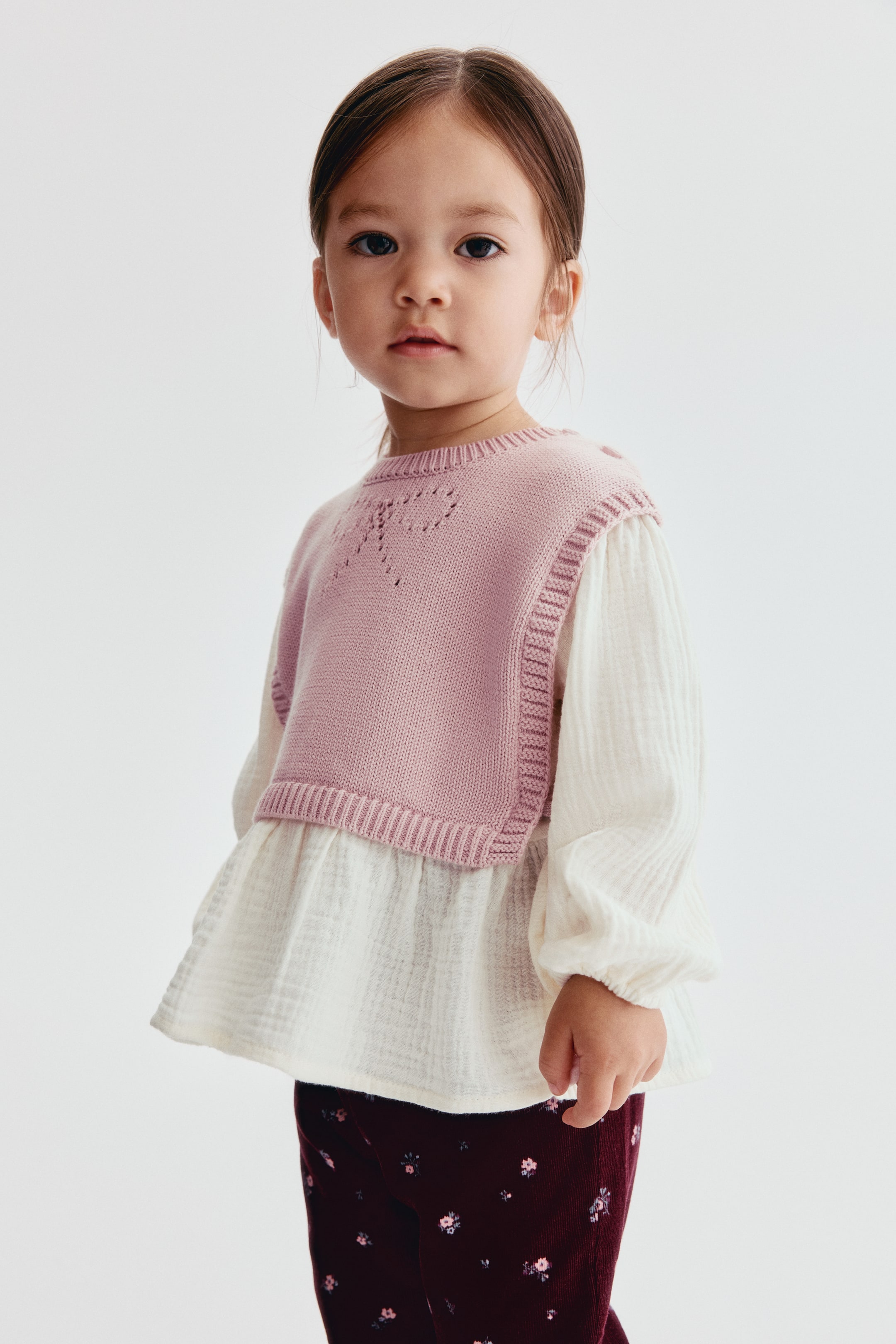 View larger image: An off-white textured cotton blouse is shown from the front, featuring a sewn-in pink knit bib with ribbed trims and a pointelle bow detail on the chest. The blouse is detailed with long puff sleeves with narrow elasticated cuffs and a flared peplum hem.