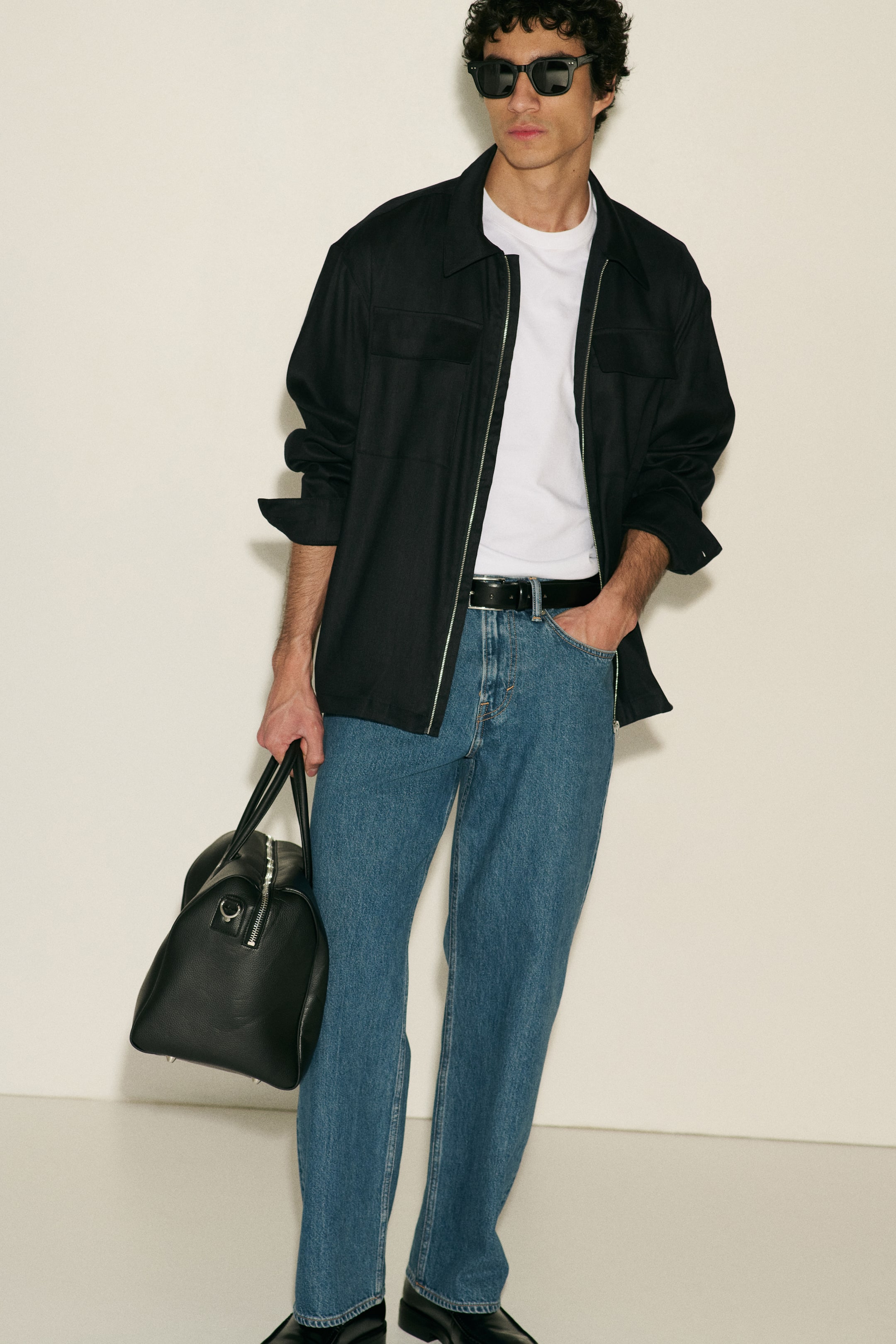 Zobacz większy obraz: Czarna torba podróżna z groszkowanej skóry jest trzymana za dwa górne uchwyty, prezentując srebrny zamek błyskawiczny. Na spodzie widoczne są ochronne metalowe nóżki.