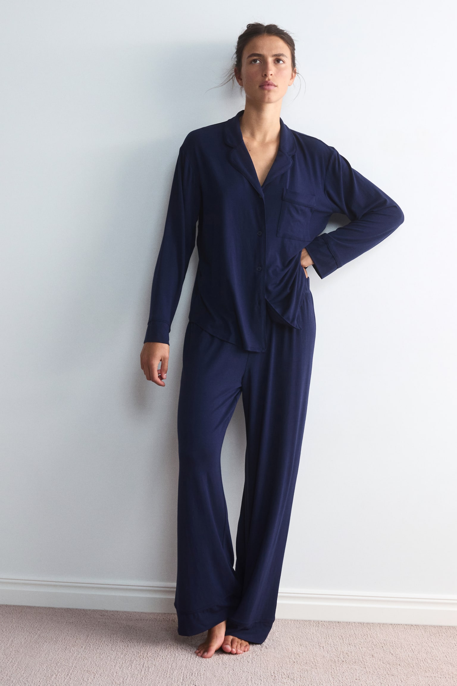 A dark blue jersey pyjama set, featuring a long-sleeved shirt with notch lapels and matching wide-leg trousers, is being worn by the model. The model is standing with one hand resting on their hip, highlighting the relaxed fit of the garments. - 1