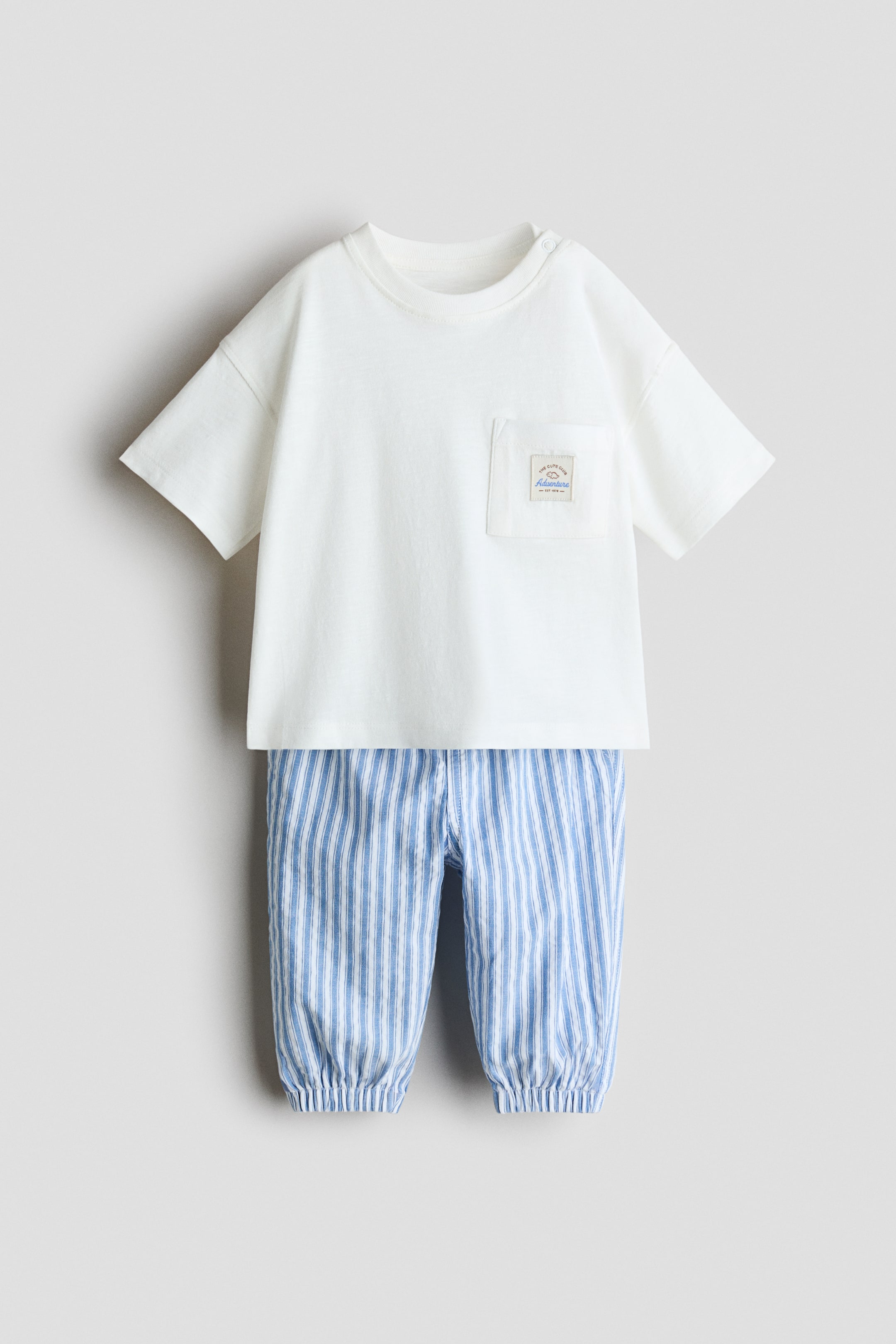 Voir une image plus grande: Ensemble en lin mélangé 2 pièces. L'ensemble présenté de face comprend un t-shirt blanc cassé à manches courtes avec un col rond, des boutons-pression sur l'épaule gauche, et une petite poche plaquée sur la poitrine gauche. Il est assorti d'un pantalon à rayures verticales bleu et blanc avec des poignets élastiqués froncés aux chevilles.