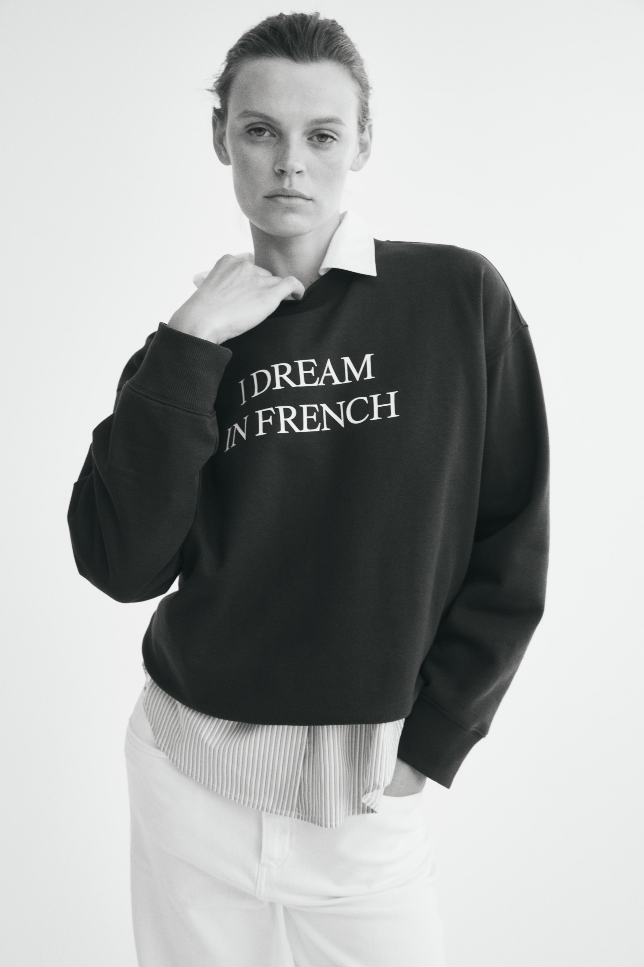 View larger image: A dark oversized sweatshirt featuring the white text "I DREAM IN FRENCH" across the chest is worn by the model. The sweatshirt is layered over a collared shirt and paired with light-colored trousers, with one hand held near the collar.