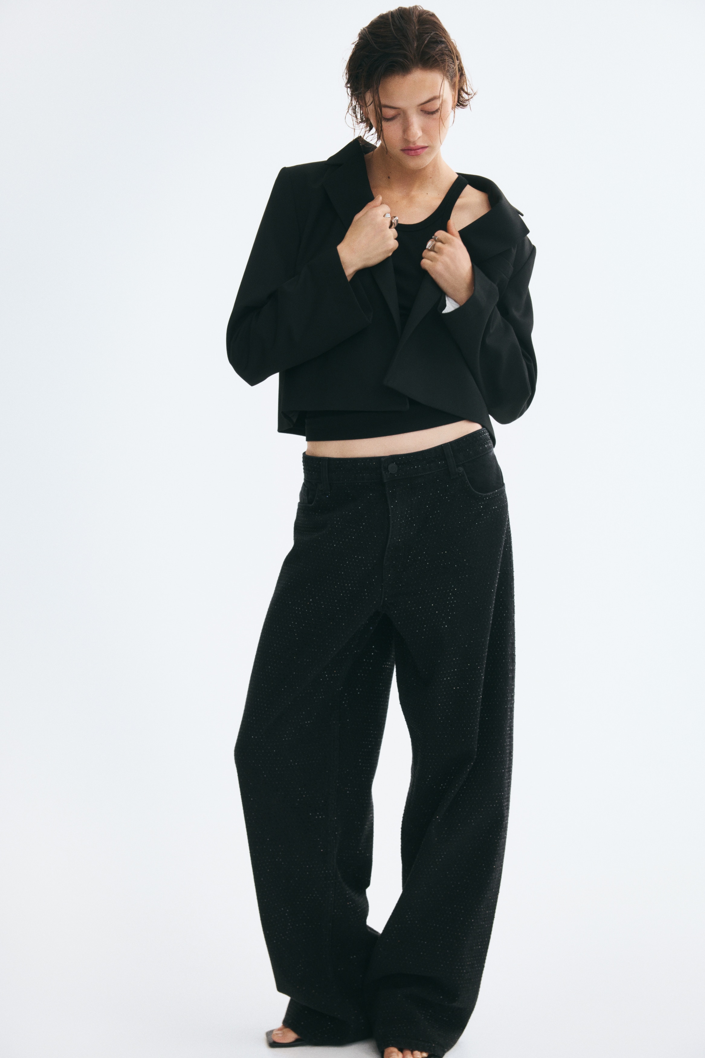 A black cropped blazer is shown front-facing, featuring notch lapels, structured shoulders, and long sleeves. The blazer is worn open with no visible closures, revealing a dark inner lining with a white rectangular label.