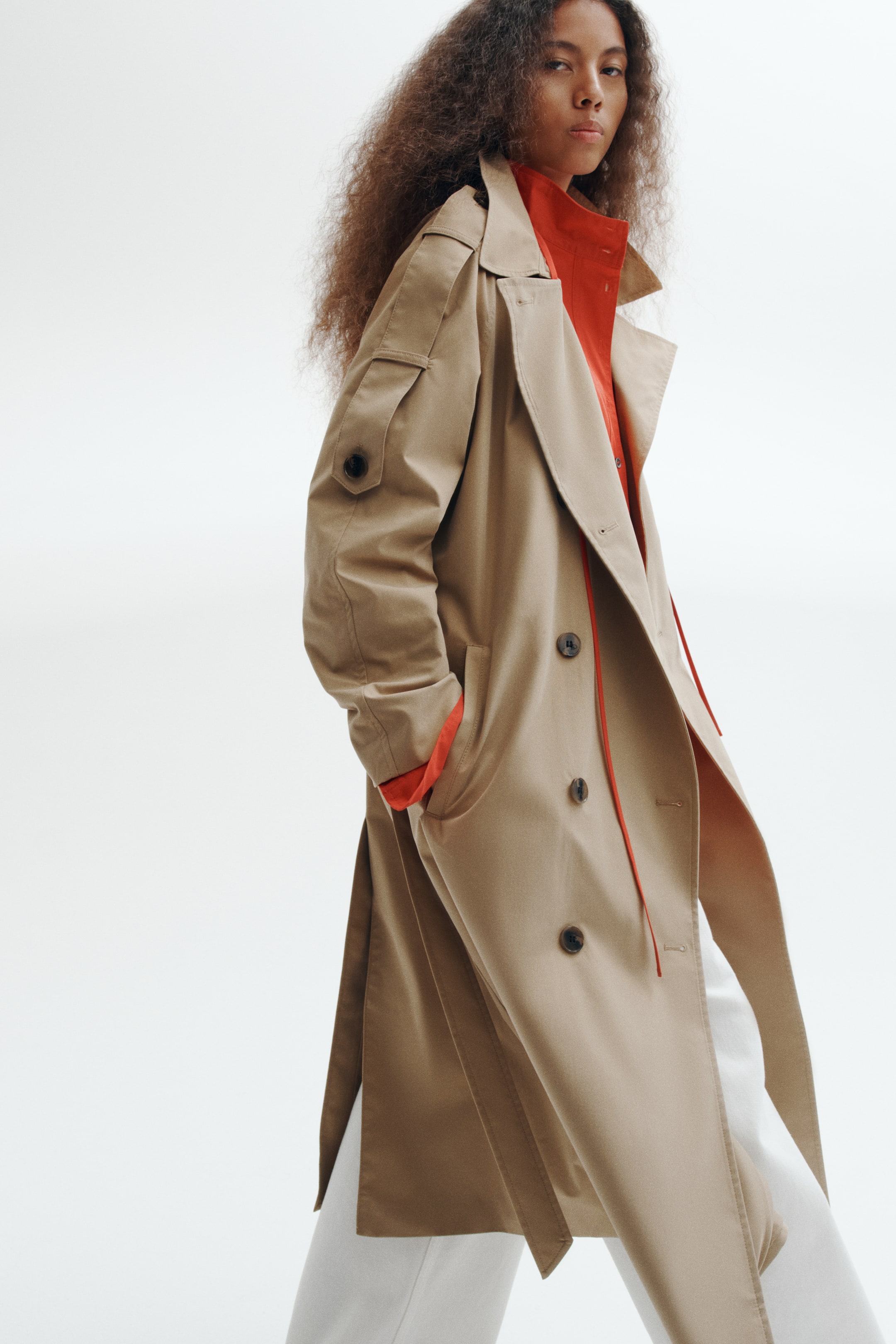Größeres Bild anzeigen: Ein beigefarbener, zweireihiger Trenchcoat wird vom Model über einem leuchtend roten Hemd und einer weißen Hose getragen. Der Oversize-Mantel verfügt über breite steigende Revers, geknöpfte Ärmelriegel und seitliche Leistentaschen, wobei das Model eine Hand in einer Tasche platziert.