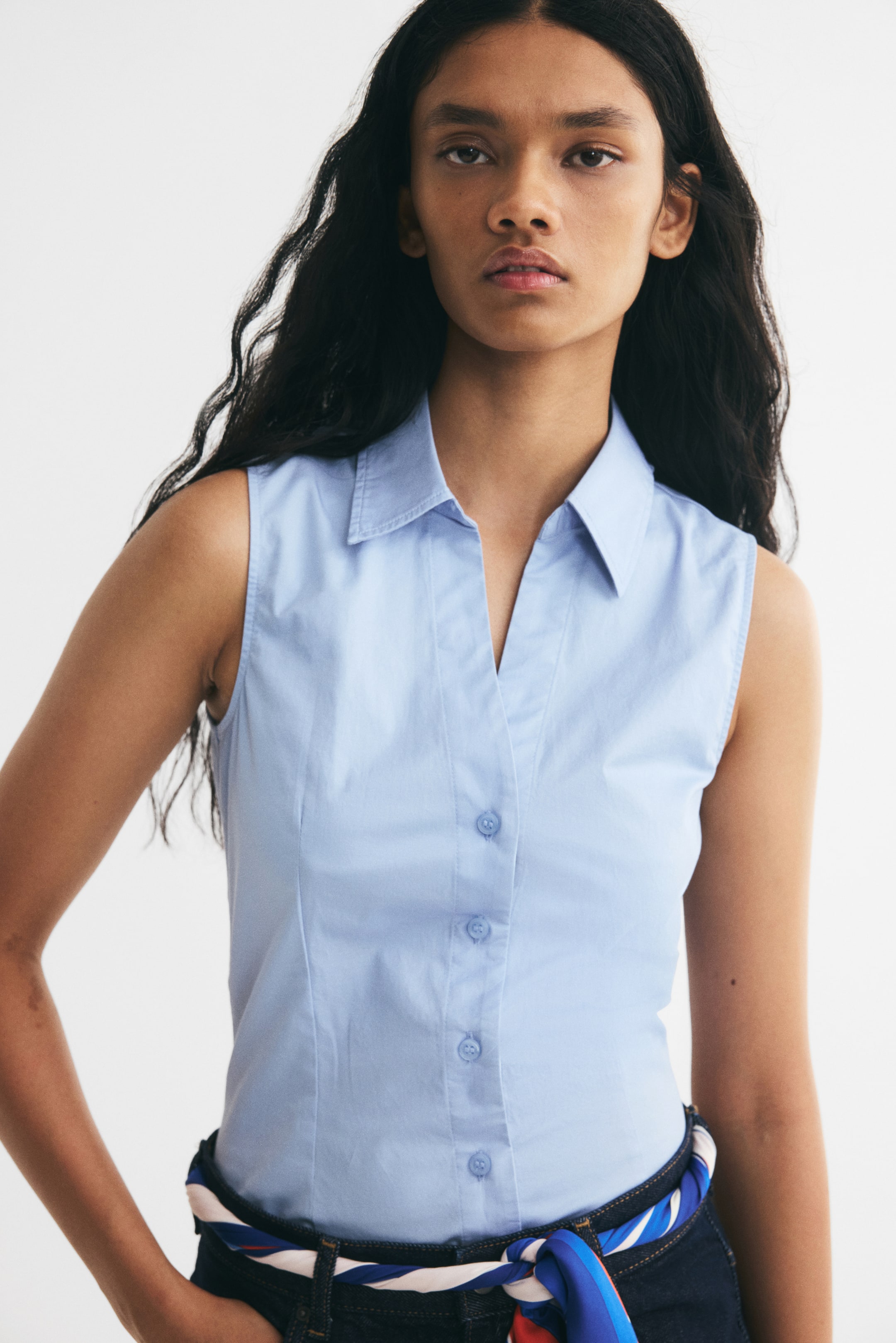 Ampliar la imagen: La modelo lleva puesta una camisa ajustada de color azul claro, sin mangas, con cuello abierto, botonadura al frente y costuras verticales de entalle. La camisa está metida dentro de unos jeans de mezclilla oscura, estilizados con un cinturón de bufanda con estampado.