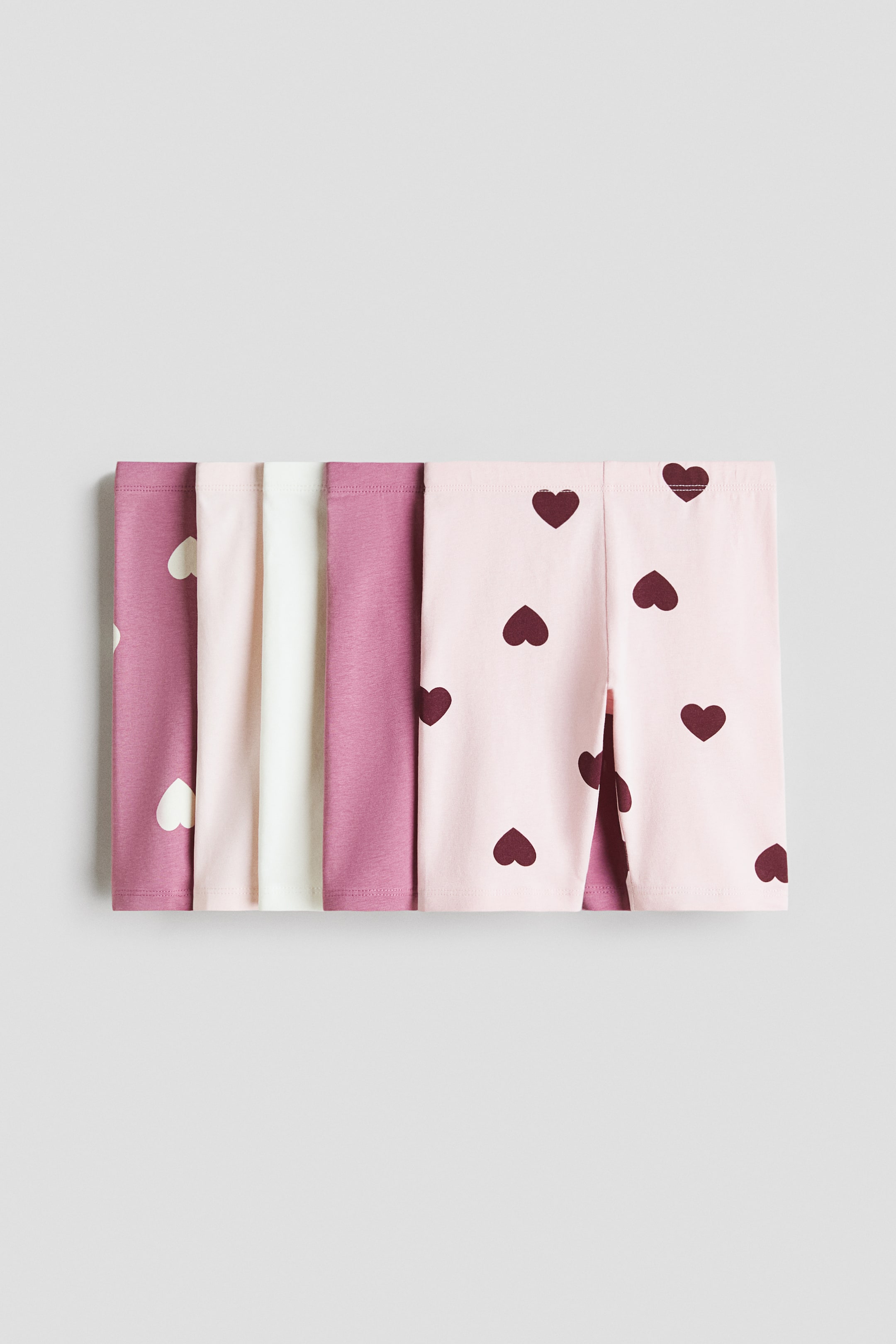 View larger image: Cycling shorts in soft cotton jersey with an elasticated waist are being displayed, folded and arranged in a horizontal row. The shorts are featuring solid colours in pink, mauve, and white, and light pink pairs with dark red heart patterns.