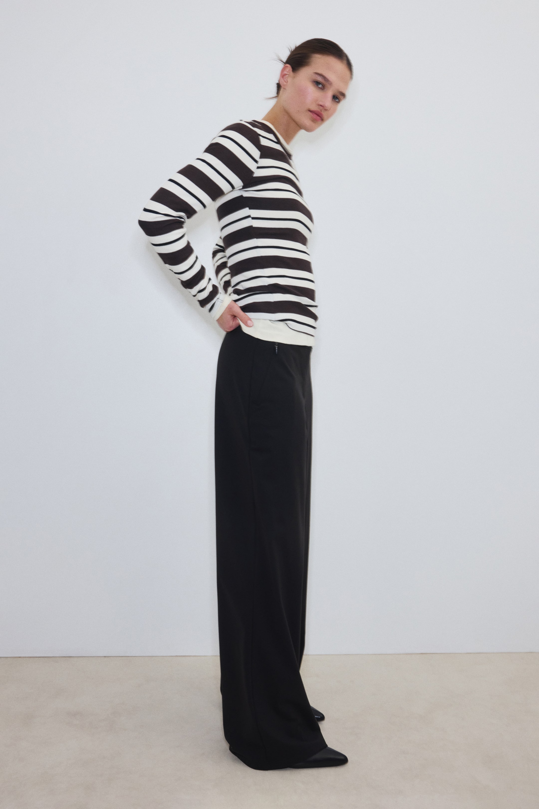 Näytä suurempi kuva: Musta leveälahkeiset housut, joissa on rento istuvuus ja joustava vyötärönauha, ovat mallin yllä. Malli seisoo, asettaen toisen käden lonkalle lähelle vyötärönauhaa, ja housut on yhdistetty raidalliseen pitkähihaiseen yläosaan ja mustiin teräväkärkisiin kenkiin.