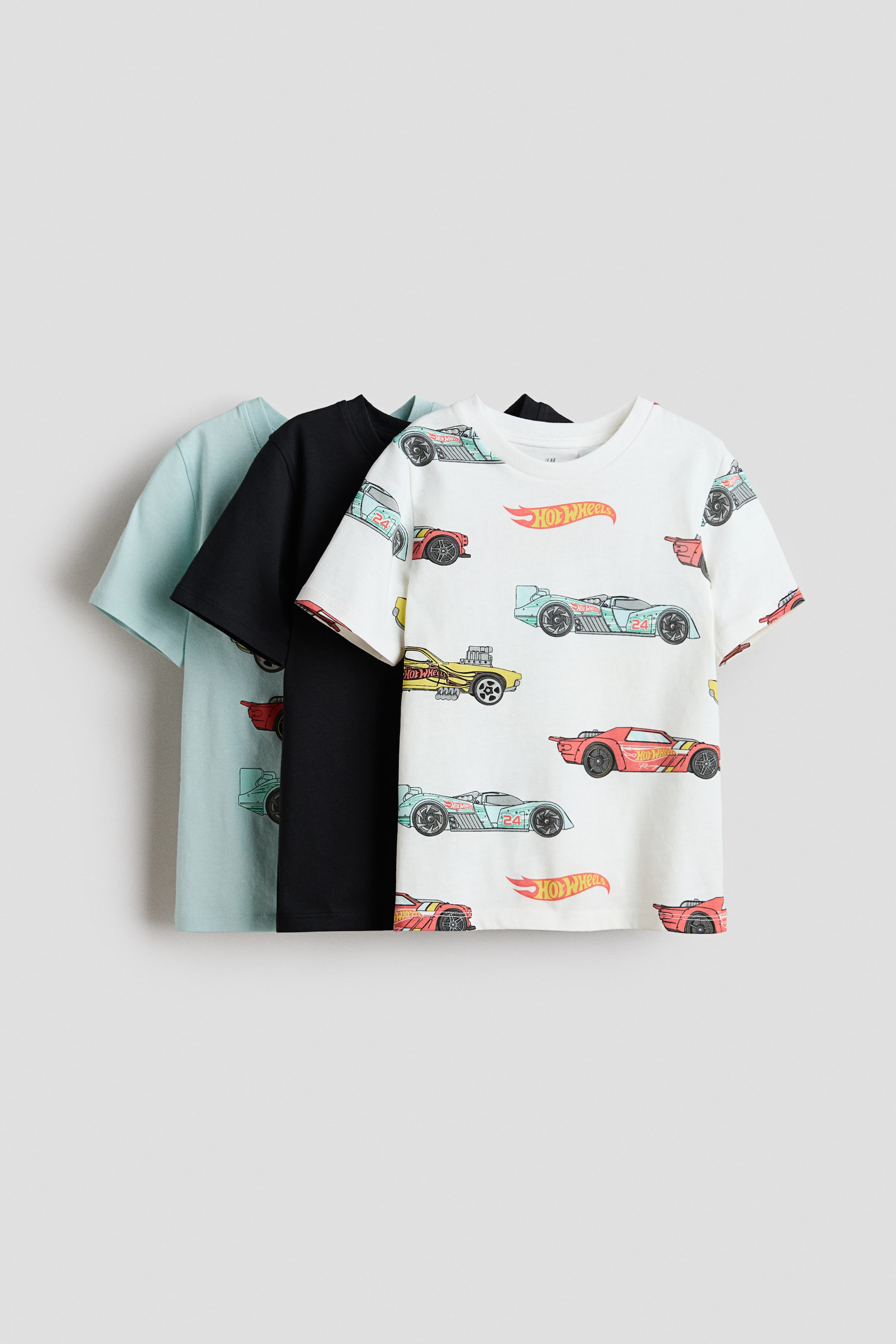 View larger image: A set of three classic t-shirts is shown from the front in a layered arrangement. The front t-shirt is white, featuring a colorful print of Hot Wheels race cars and logos and a rib-trimmed crew neckline, while a plain black t-shirt and a plain light blue t-shirt are partially visible behind it, both also displaying rib-trimmed necklines.