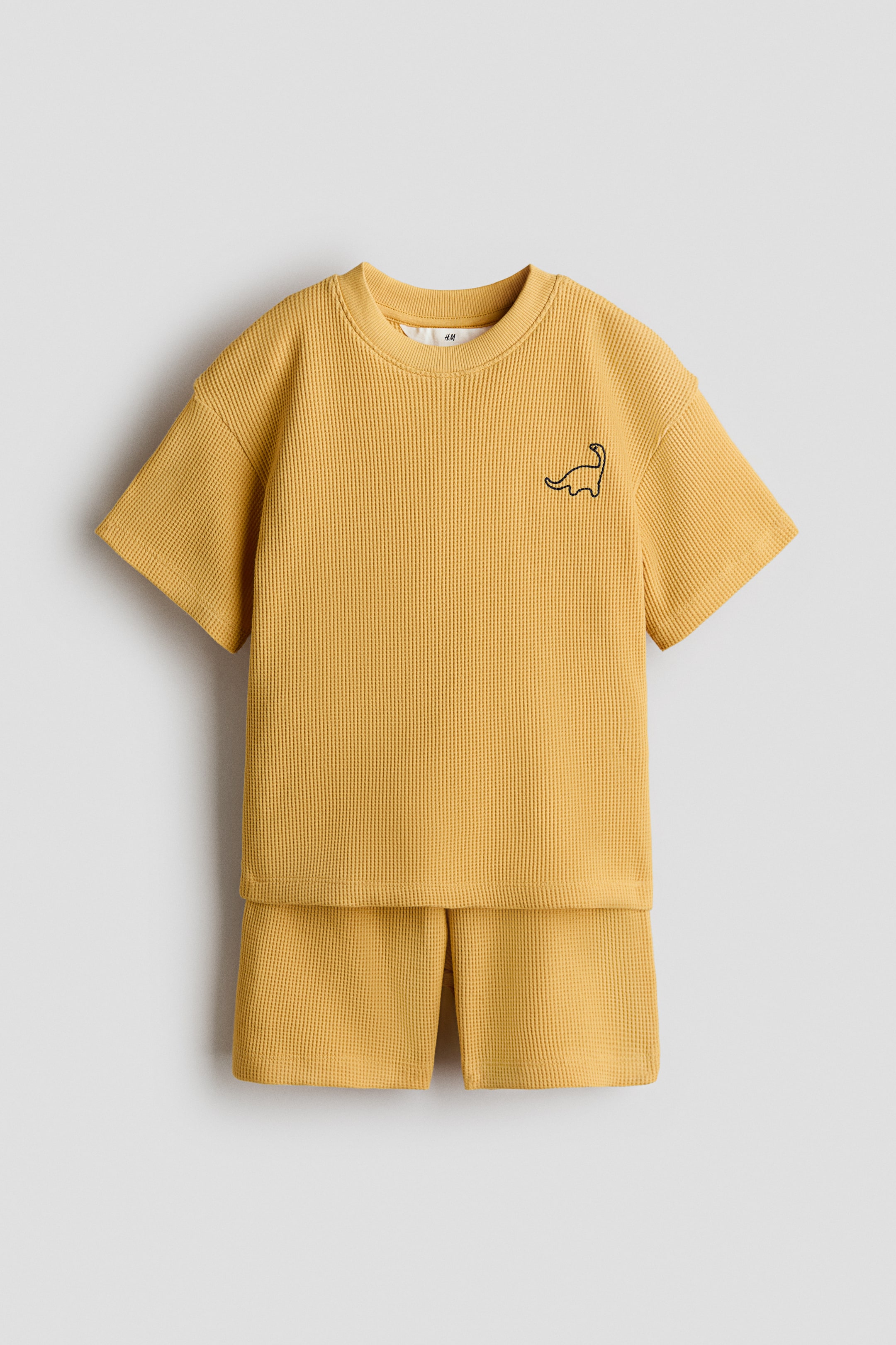 View larger image: A mustard-yellow waffle-knit T-shirt and matching shorts set is displayed facing forward. The short-sleeved T-shirt features a ribbed crew neck and a black embroidered dinosaur outline on the left chest.
