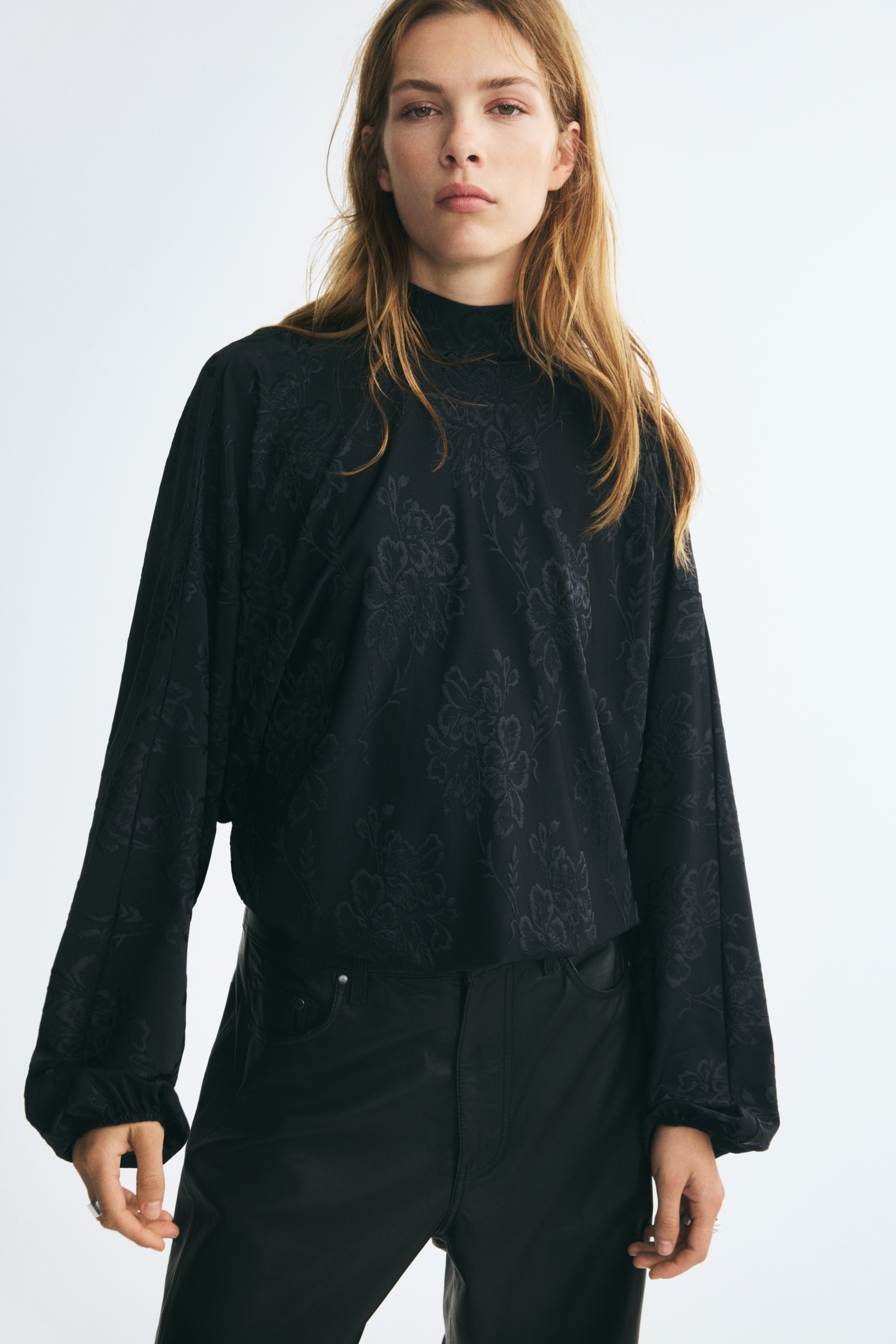 A black, oversized blouse in tonal floral jacquard fabric is shown from the front, featuring a high stand collar and voluminous balloon sleeves with elasticated cuffs. The hem of the blouse gathers into an elasticated bubble hem, creating a puffy silhouette.