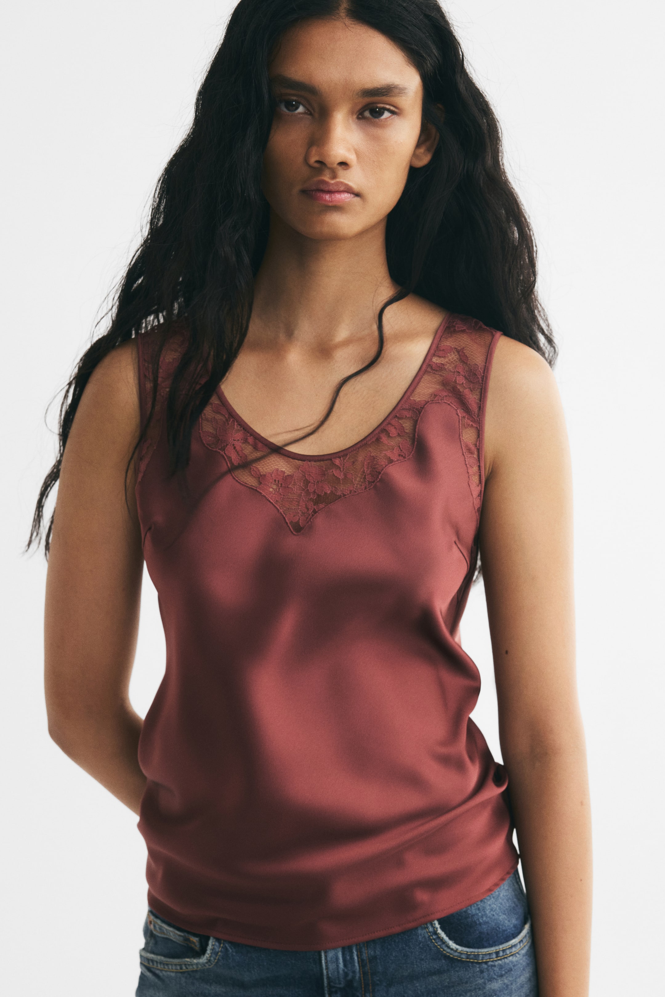 View larger image: The model is wearing a dark rose satin top with a scoop neckline, delicate lace inset, and wide shoulder straps, styled with blue denim jeans.