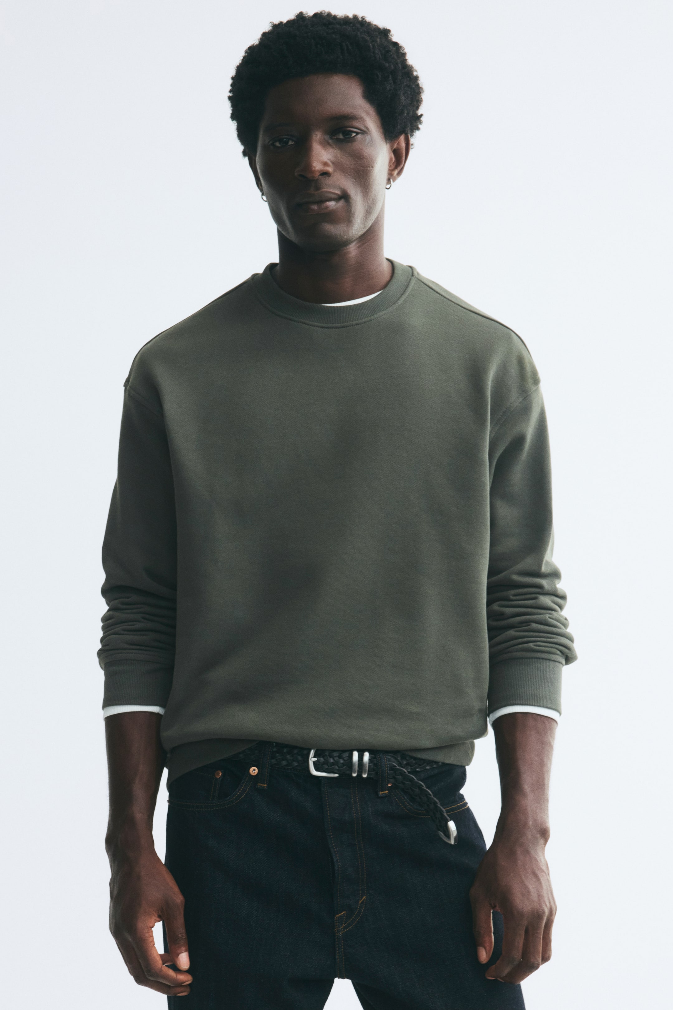 View larger image: An olive green crewneck sweatshirt in a relaxed fit is being worn by the model, featuring dropped shoulders and ribbed trims at the neckline, cuffs, and hem. The sweatshirt is layered over a white tee and paired with dark denim jeans.
