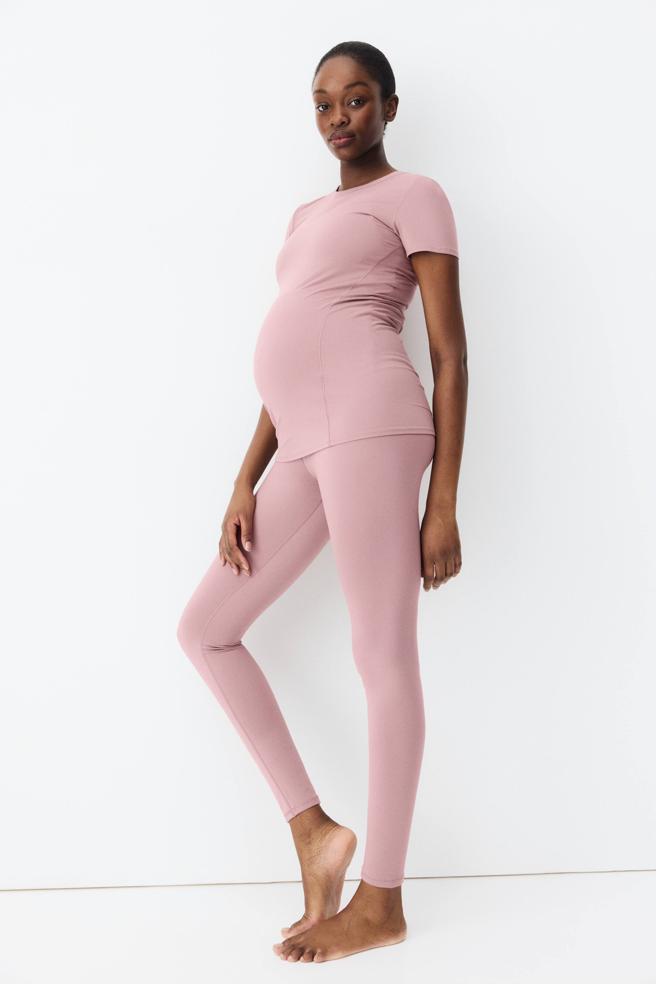 Ampliar la imagen: Se está mostrando un conjunto de ropa activa de maternidad en color rosa polvoriento, que cuenta con una camiseta de manga corta y cuello redondo con costuras laterales que se recogen sobre el vientre, y leggings a juego de longitud completa. El conjunto ajustado se presenta desde un ángulo ligeramente lateral.