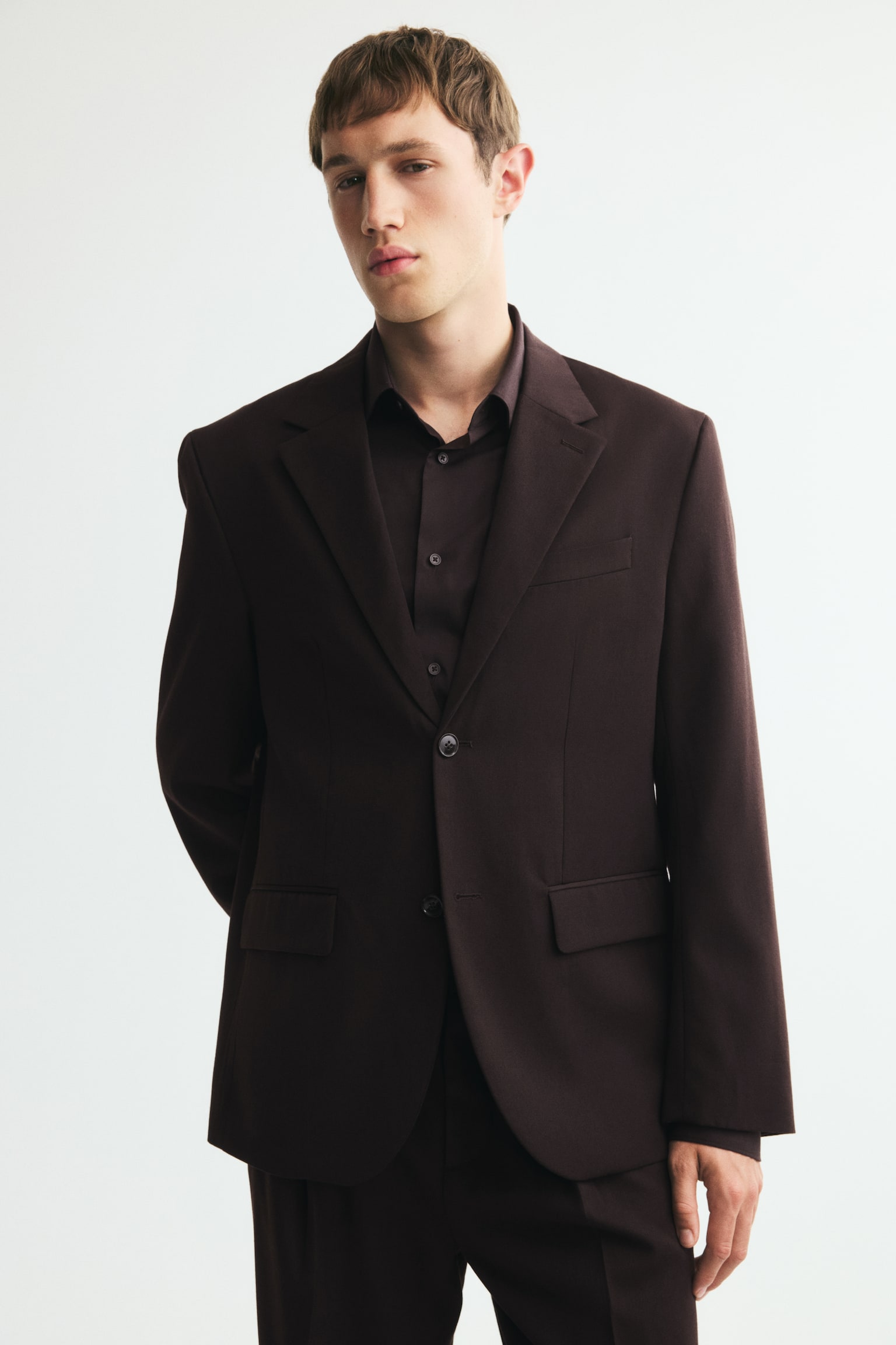 En mørkebrun blazer præsenteres forfra, med notch-revers og en to-knaps lukning. Den viser to klaplommer ved hofterne, en enkelt paspoleret brystlomme og manchetter med knapper, med sort foring synlig ved kraven. - 1