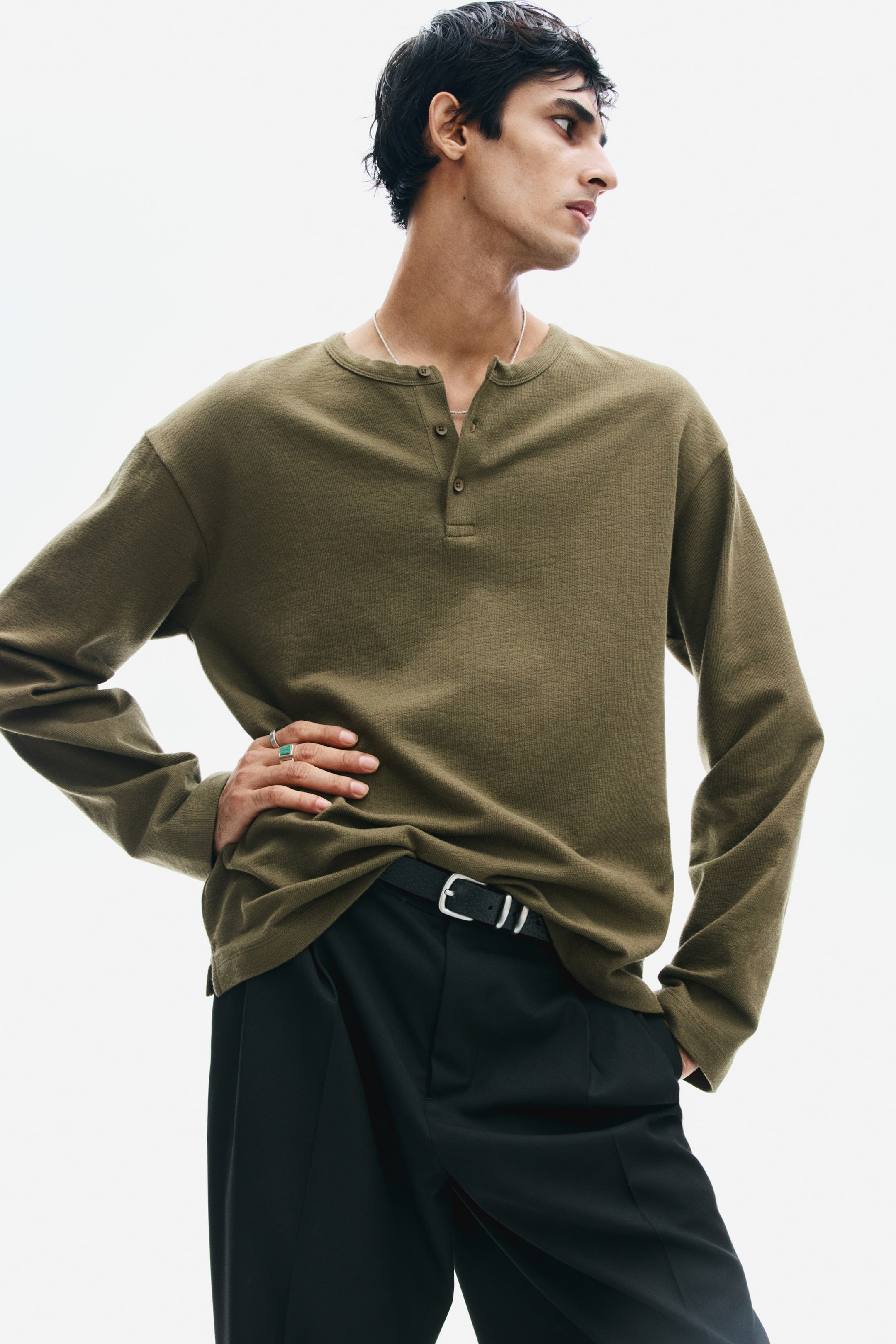 View larger image: The model is wearing a long-sleeve henley shirt in olive green, featuring textured knit fabric and a partially unbuttoned collar. The relaxed-fit shirt is styled slightly tucked into black trousers with a black belt.