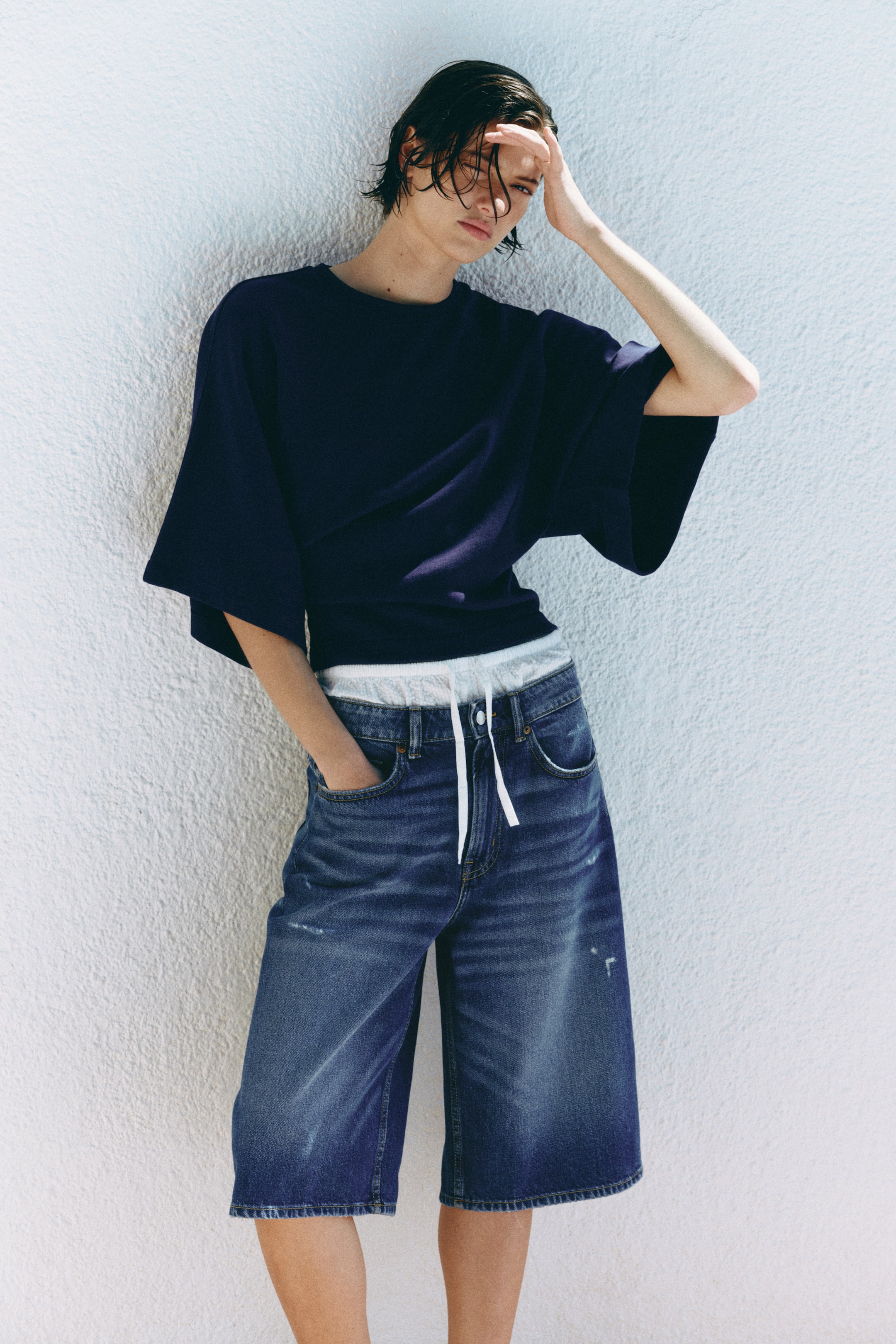 View larger image: The navy blue, loose-fitting top with wide short sleeves and a round neck is being worn, revealing a white elasticated undershirt waistband with a drawstring. Distressed blue denim shorts, featuring front pockets and a matching white drawstring, are completing the outfit, extending to just above the knees.