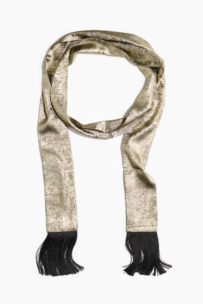 A metallic gold scarf with a textured, shimmering finish is draped in a loose loop, facing front with its two ends hanging straight down. Each end of the scarf is finished with long, black tassel fringe. - 1
