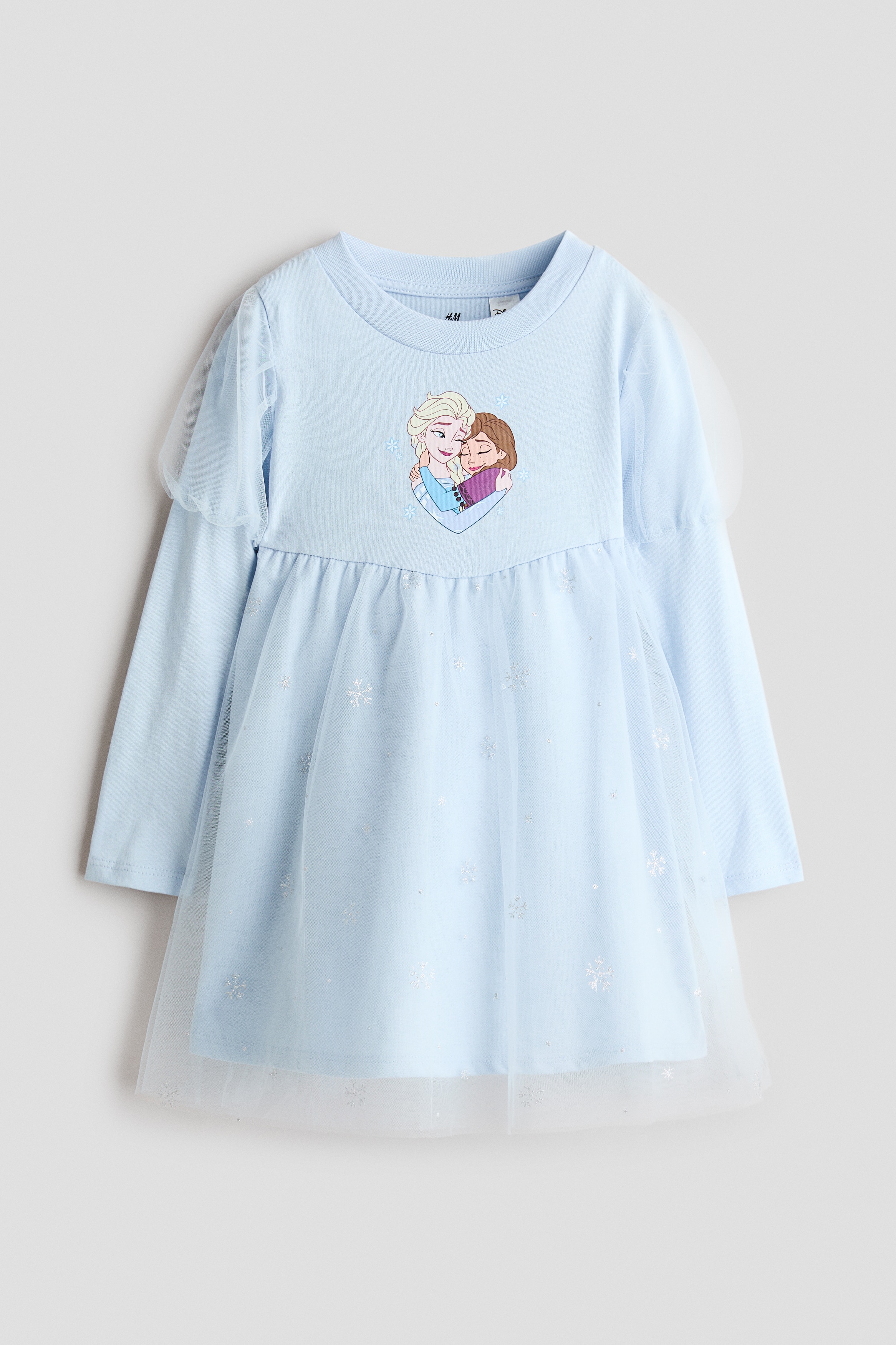 The model is wearing a light blue long-sleeved dress featuring a printed character motif on the front chest and a flared skirt with a sheer tulle overlay in a snowflake pattern. The dress has a round neckline, gathering at the waist, and long sleeves layered with short, puffed tulle sleeves at the shoulders.