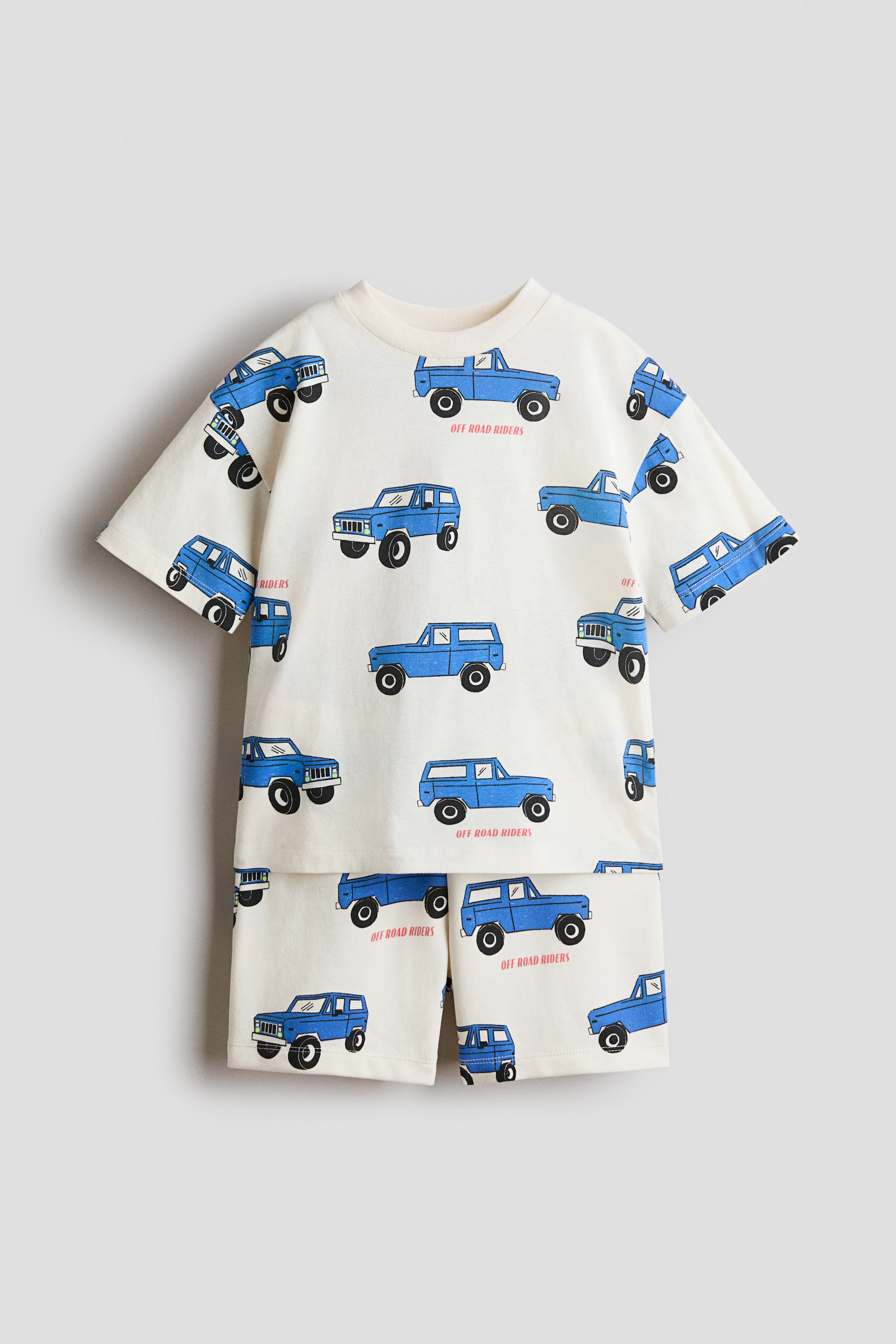 View larger image: A cream-coloured short-sleeved T-shirt and matching shorts set, shown from the front, features a repeating pattern of blue off-road vehicles with the text "OFF ROAD RIDERS" printed beneath some of them.