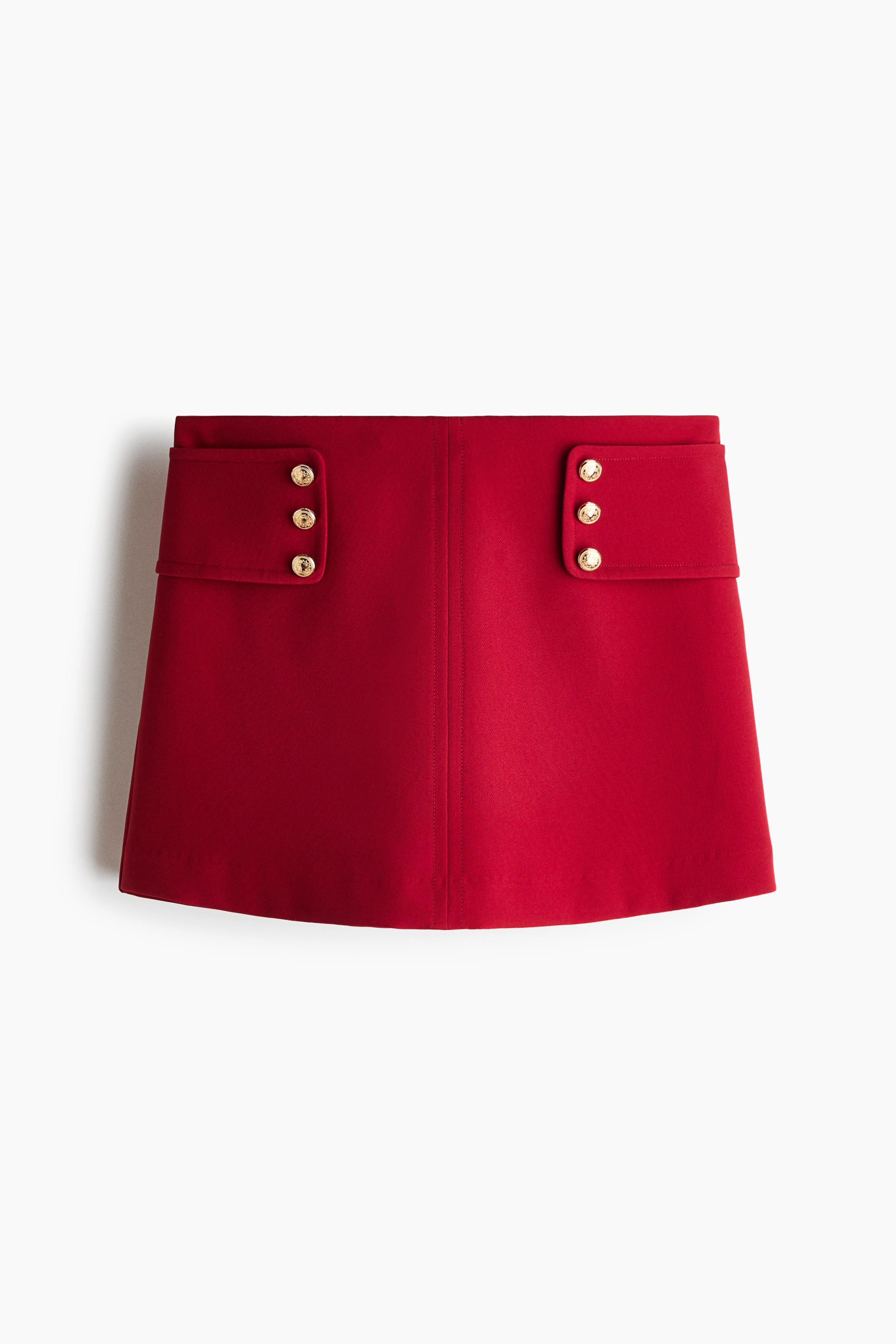 La modelo lleva puesto un skort corto rojo con una costura vertical al frente y trabillas anchas en la cintura con botones decorativos de tono dorado. El skort se combina con una playera sin mangas blanca de canalé.