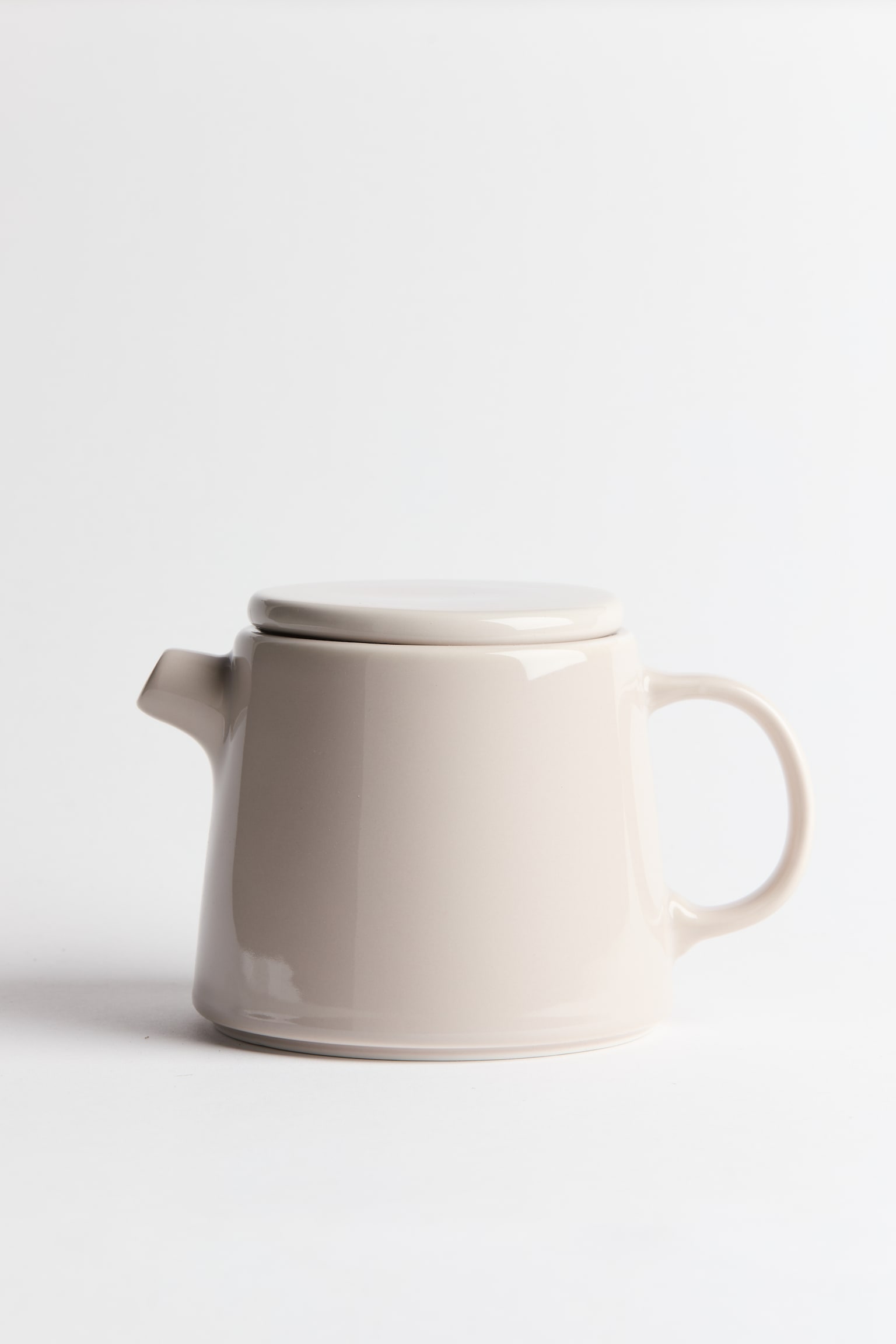 A porcelain teapot with a glossy, light beige glaze is shown in side profile, showcasing its slightly tapered cylindrical body. The teapot features a short, angled spout on the left, a rounded loop handle on the right, and a flat matching lid resting on top.