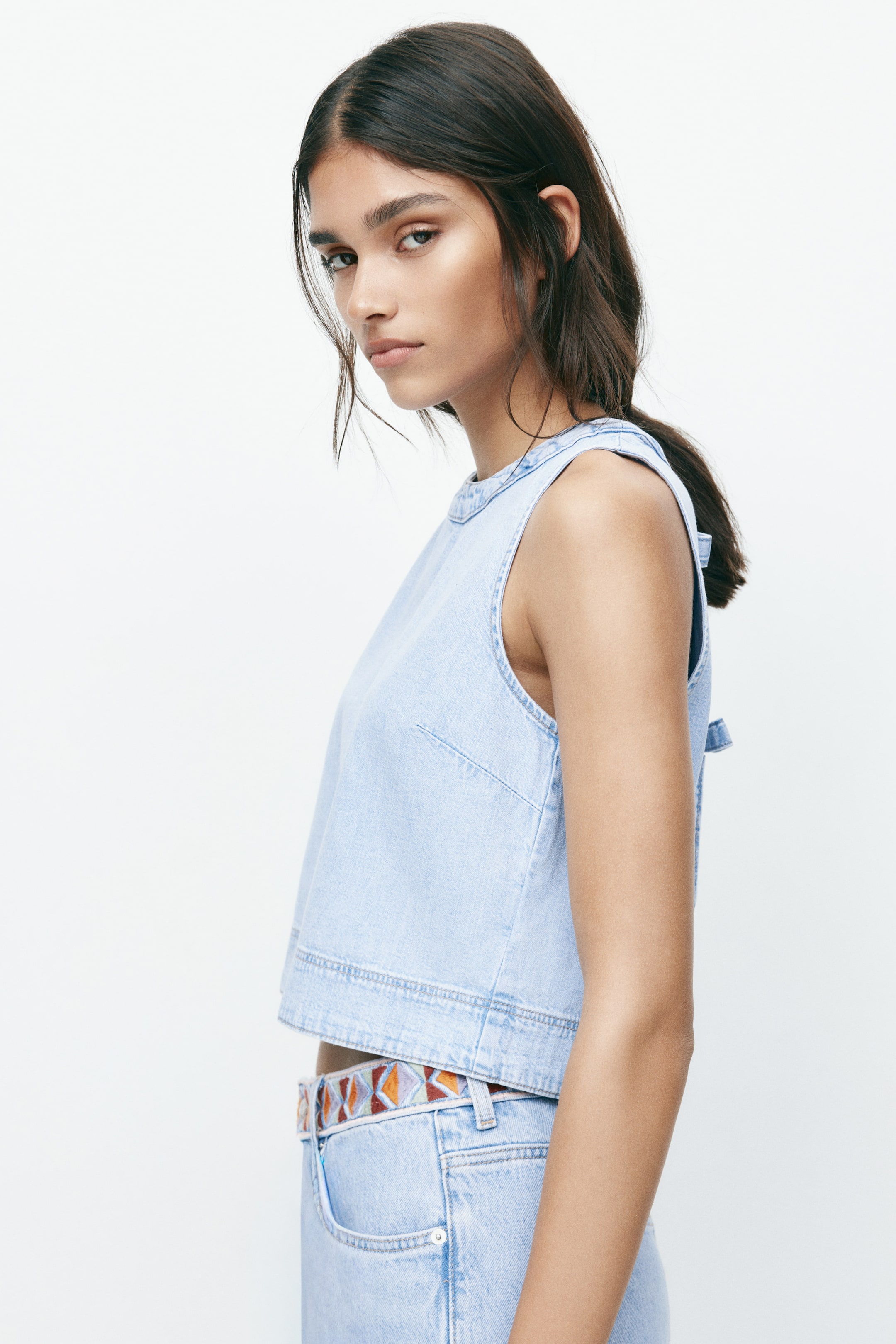 Größeres Bild anzeigen: Ein hellblaues Denim-Crop-Top wird aus einem seitlich-vorderen Winkel betrachtet und verfügt über einen Rundhalsausschnitt und ein ärmelloses Design. Es zeigt sichtbare Paneelnähte an der Vorderseite, einen geraden Saum und ein kleines Bindedetail an der Rückenschulter.