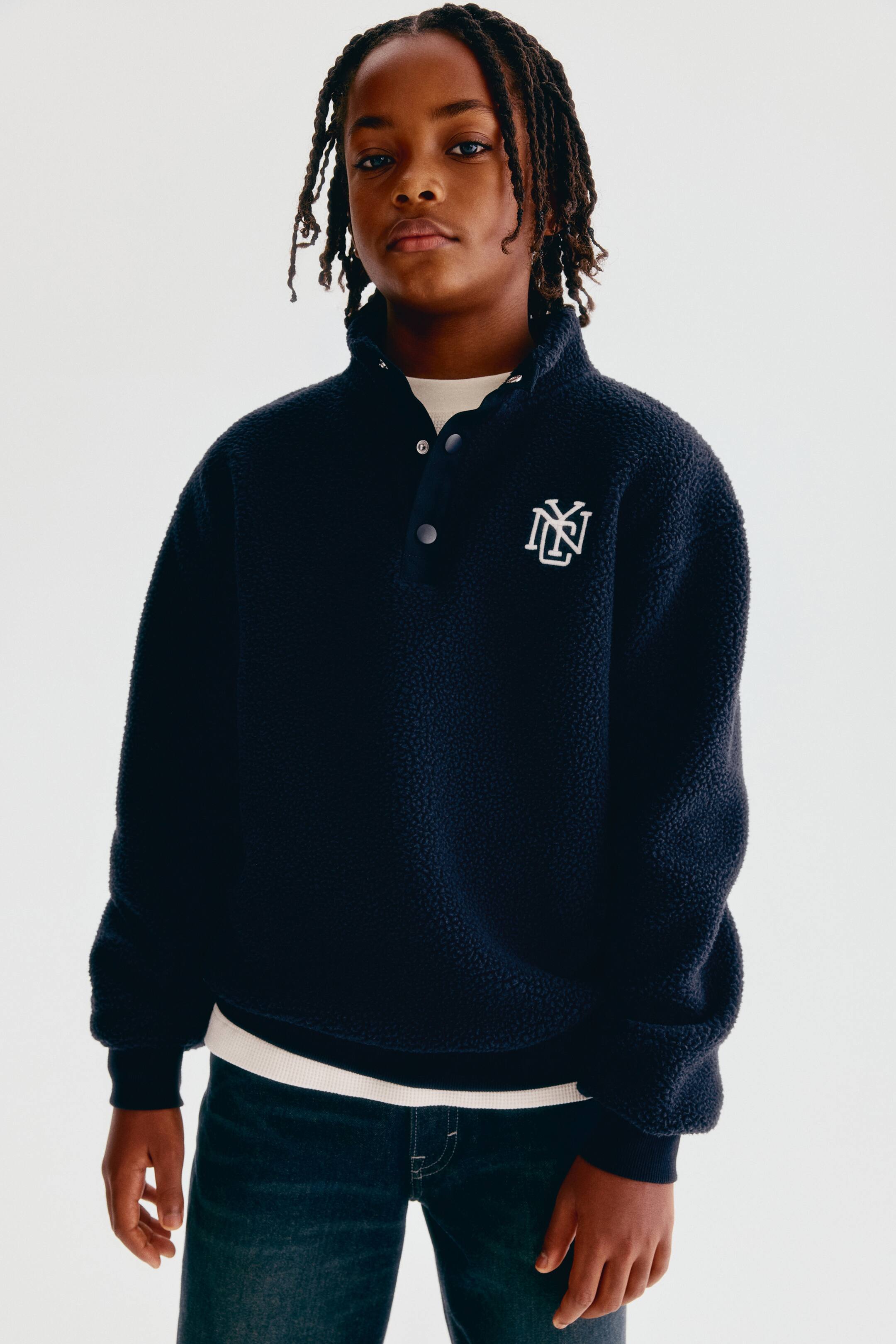 View larger image: A navy blue teddy sweater is shown from the front, featuring a stand collar with a press-stud placket and a white embroidered monogram on the chest. The sweater has dropped shoulders, ribbed cuffs, and a ribbed hem, with a layered white T-shirt peeking out at the collar and hem.