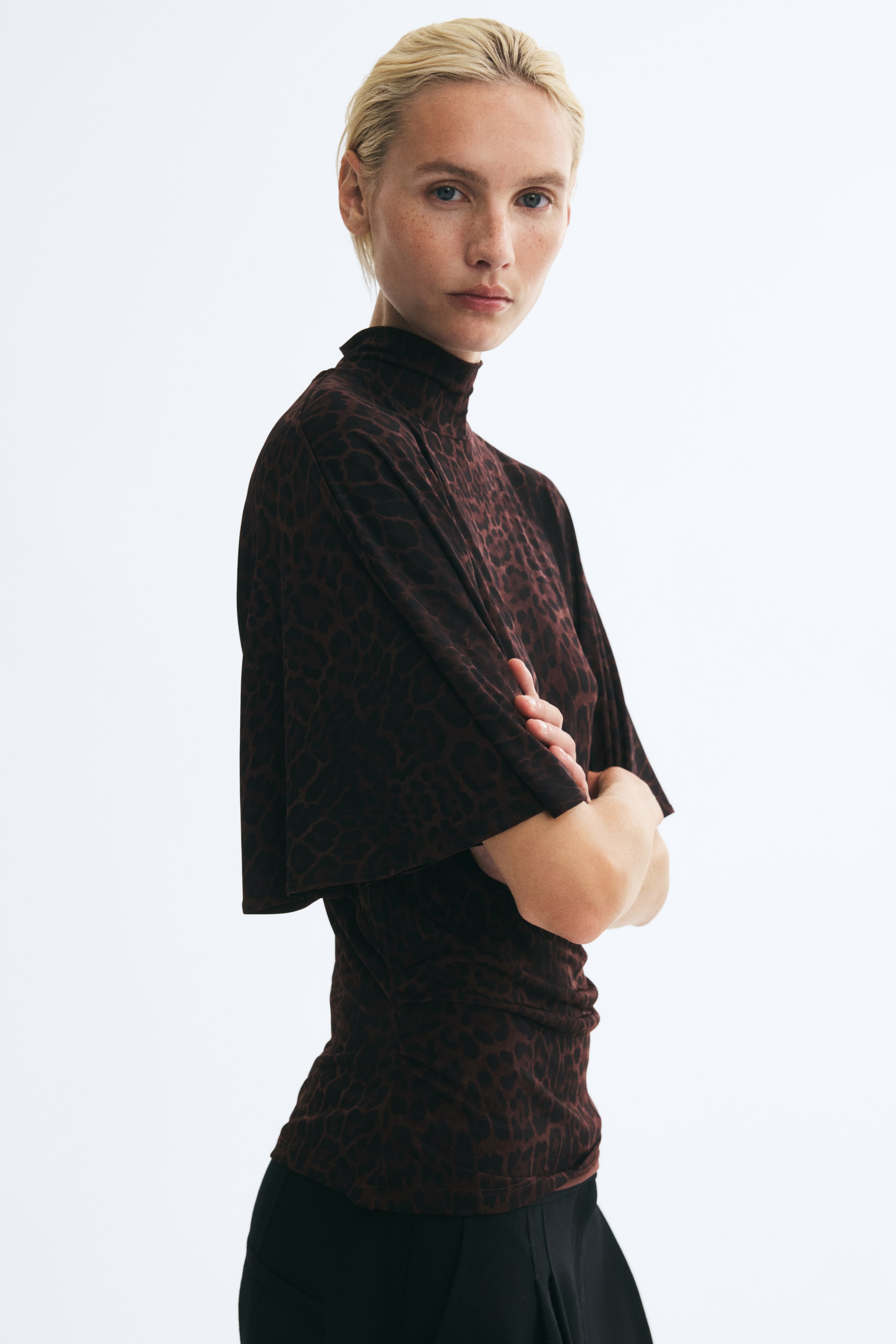 Se større billede: En top i elastisk jersey med leopardprint, høj hals og korte, draperede batwingærmer bæres af modellen, der står med korslagte arme. Den løstsiddende top styles med sorte bukser.