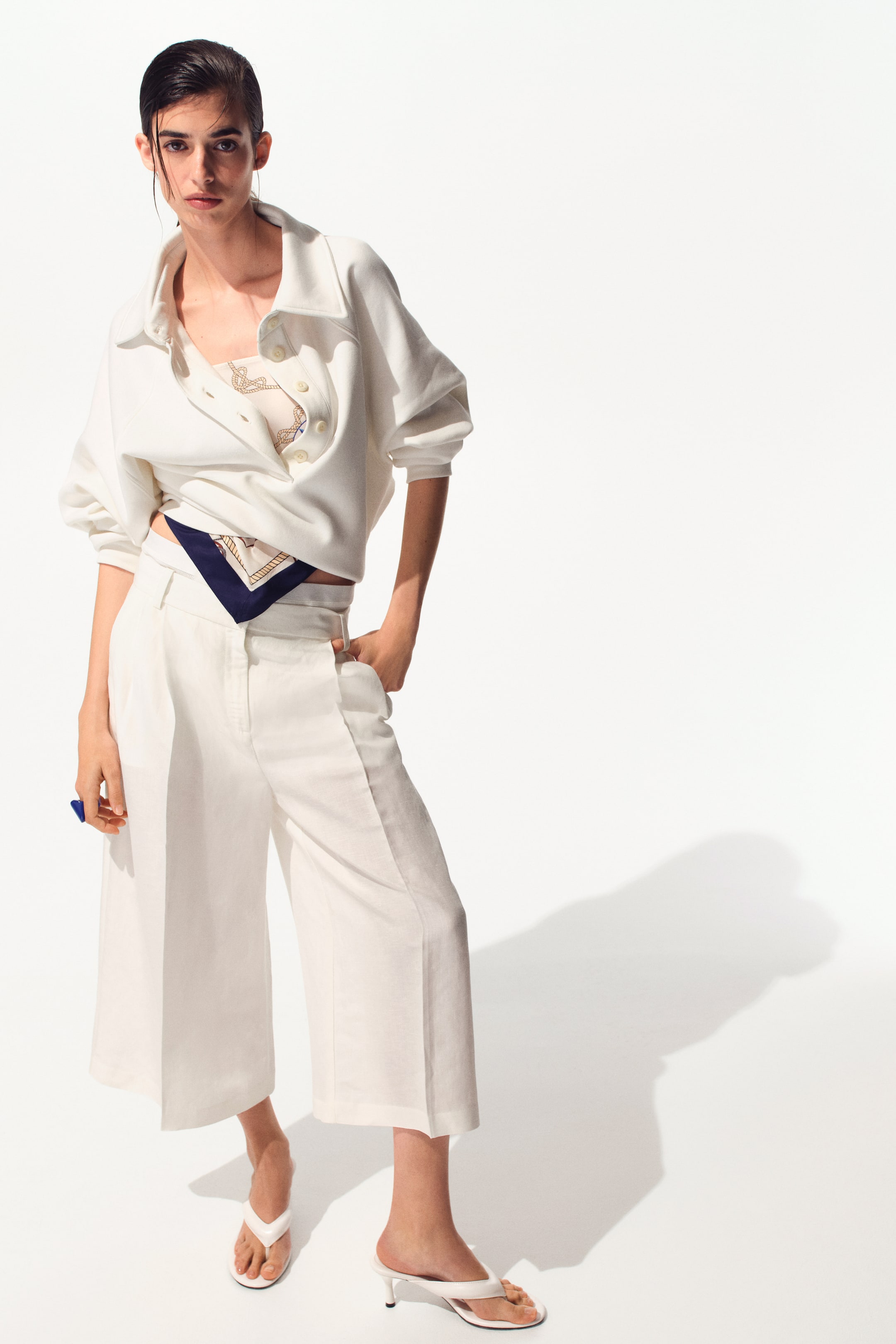 Visualizza immagine più grande: L'outfit presenta una felpa bianca cropped con colletto e maniche raglan, con un'abbottonatura parziale, sovrapposta a un top a fascia color crema con motivo. I pantaloni bianchi a gamba larga e taglio cropped completano l'insieme, con una sciarpa a motivo blu scuro infilata nella cintura e sandali infradito bianchi con tacco basso.