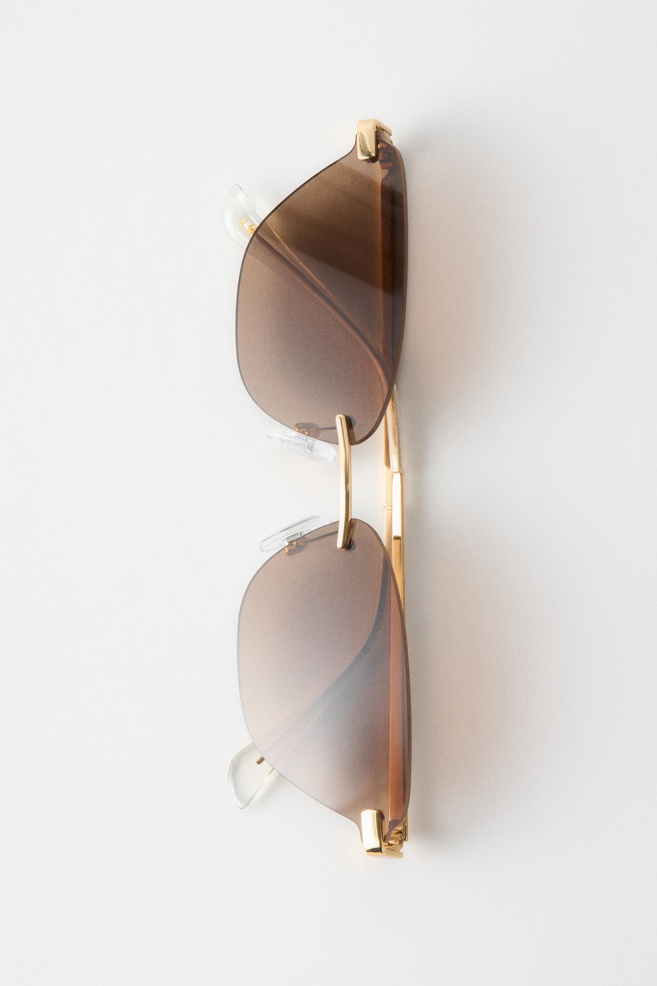Voir une image plus grande: Des lunettes de soleil ovales sans monture, à forme légèrement géométrique, sont montrées de face, avec des verres teintés dégradés bruns, un pont en métal doré et des plaquettes nasales réglables transparentes. Les branches en métal doré sont repliées derrière les verres.