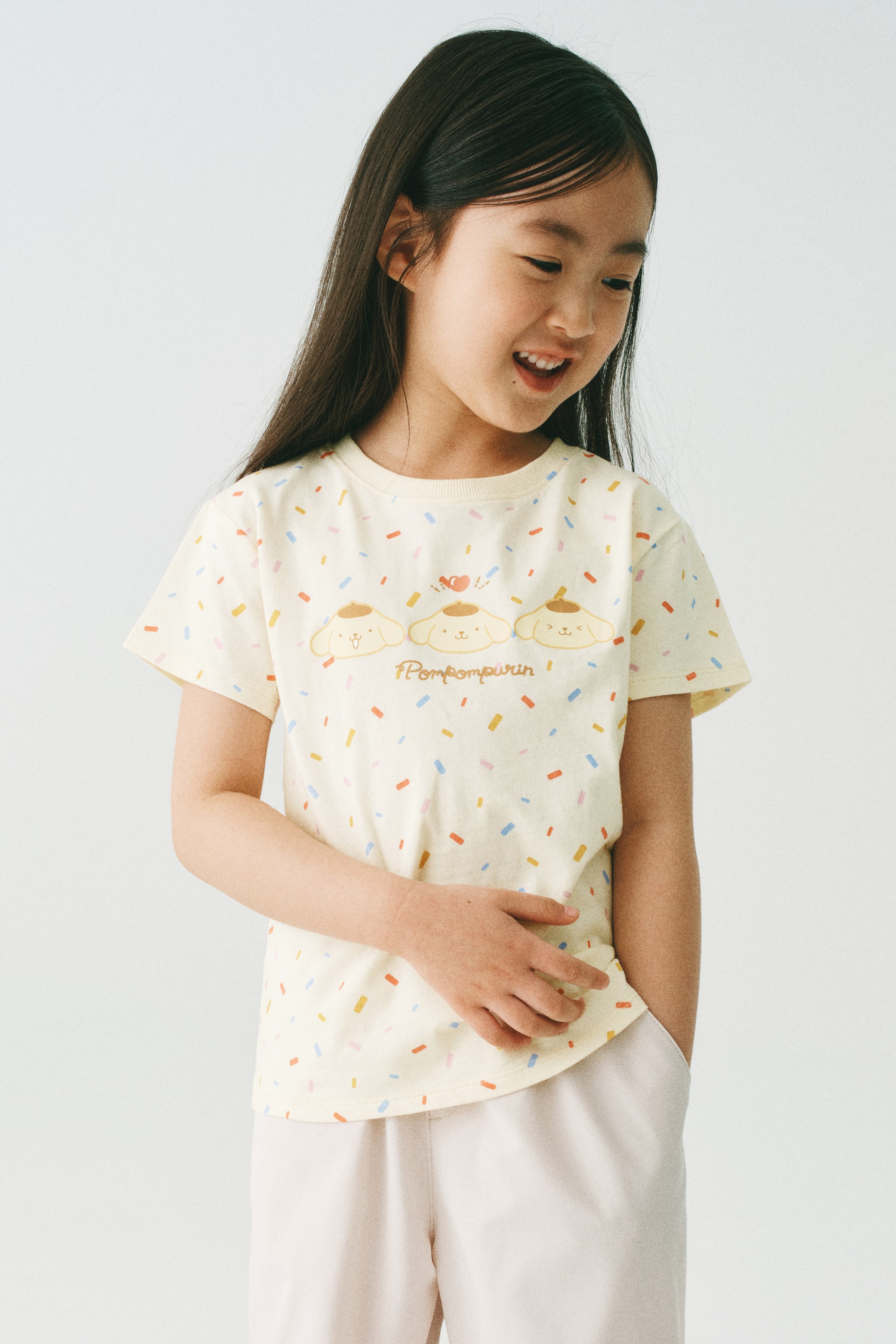 View larger image: The model is wearing a T-shirt made of soft, printed cotton jersey, featuring a ribbed trim around the neckline. This light yellow T-shirt showcases a vibrant confetti pattern and Pompompurin characters on the front. The model is seen with a hand placed on the front of the shirt.